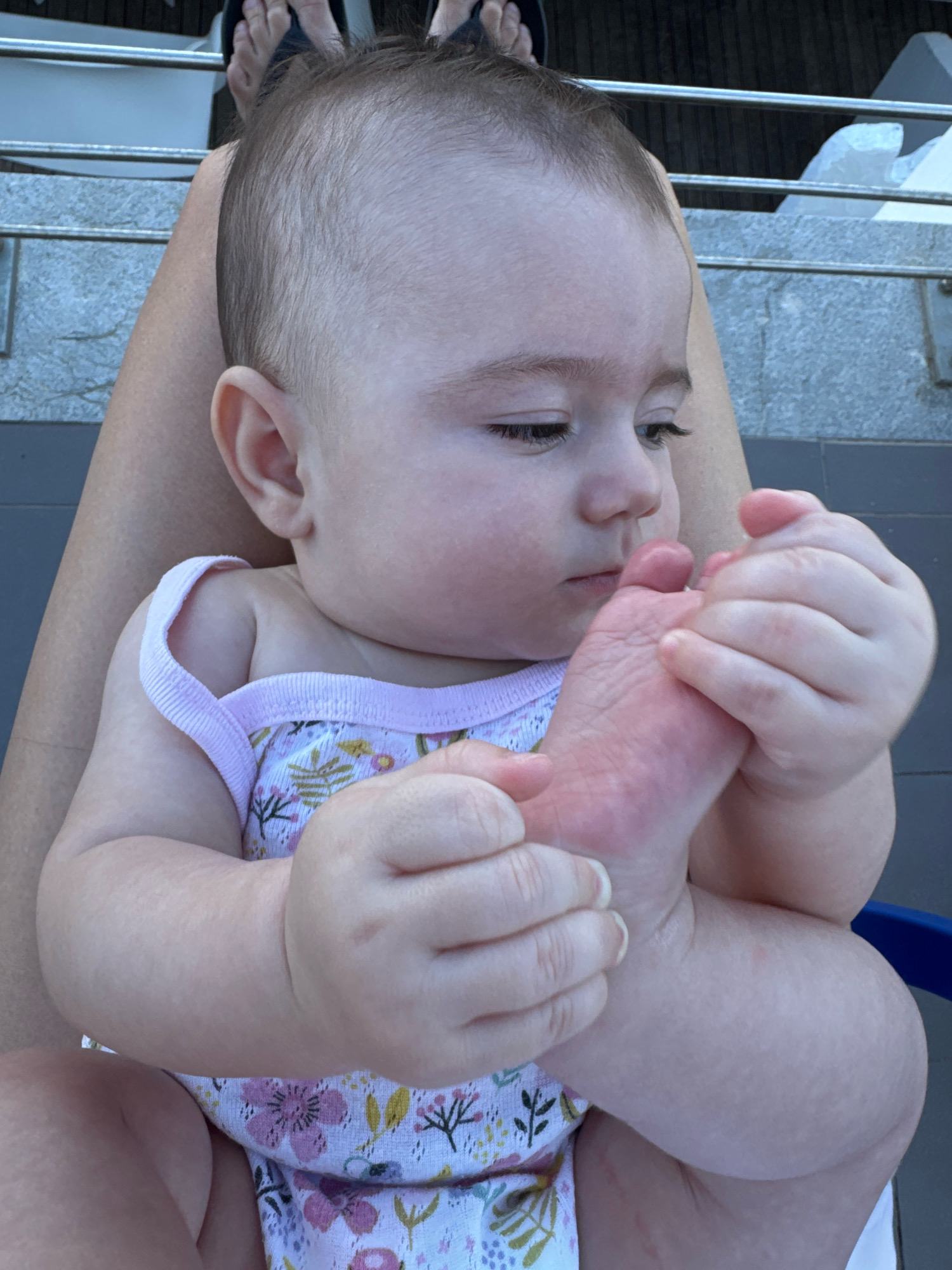 The image shows a close-up of a baby sitting on someone's lap. The baby is looking down at its foot, which it is holding with both hands. The baby is wearing a floral-patterned onesie. The background is out of focus, but appears to be an outdoor setting with some metal railings and steps. The image does not provide any specific information about the location in Isola delle Femmine, Italy.