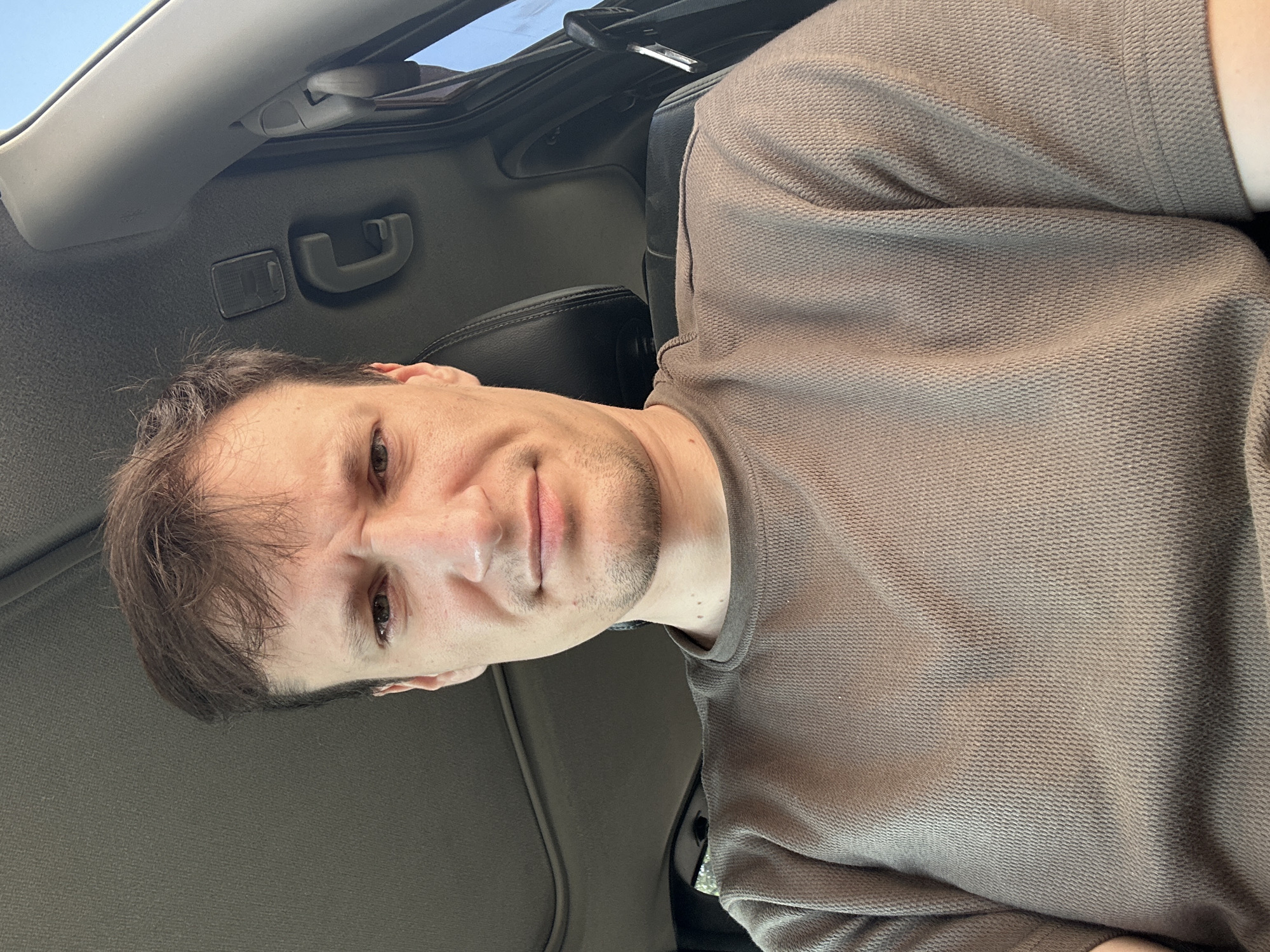 A man with short, brown hair and a light complexion is shown from the chest up, sitting in the driver's seat of a car. He has a confused or displeased expression on his face. The interior of the car is visible, including the ceiling, a sunroof, and the headrest of the passenger seat. The background is out of focus and appears to be an outdoor setting, possibly a city or town. The man is wearing a brown, short-sleeved shirt. There is no indication of the specific location within Shatava, Ukraine.
