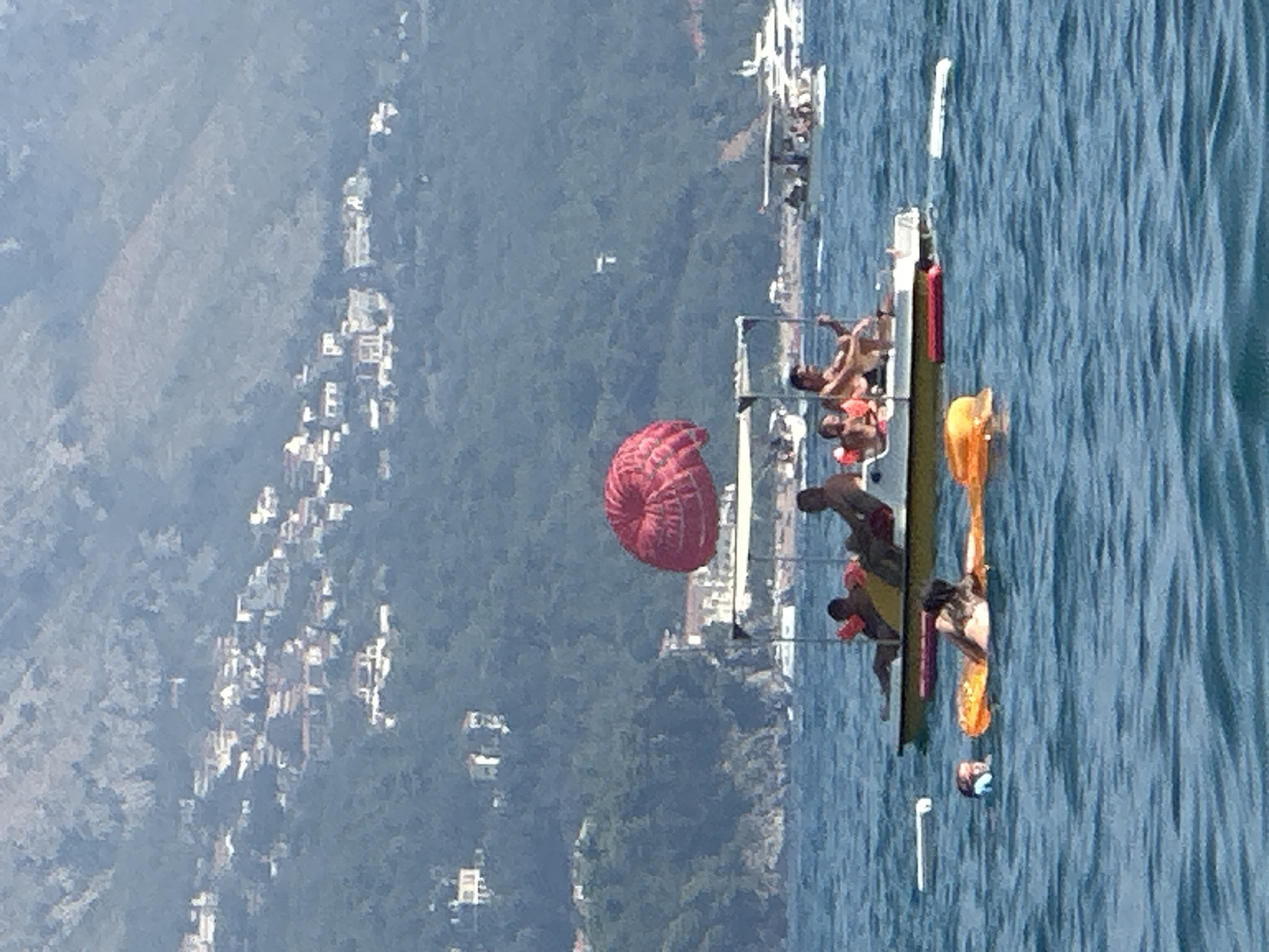 The image shows a body of water with several people engaged in various water activities. In the foreground, a person is floating on an orange inflatable, while another person in the water wears a blue swim cap and goggles. Behind them, a white pedal boat is occupied by at least four individuals. A large, red parasail is deployed above the pedal boat, suggesting a parasailing activity. In the background, a densely populated hillside is visible, dotted with white buildings. This hillside is characteristic of the city of Boreti, Montenegro, which is known for its coastal location and the proliferation of structures on its slopes. Further in the background, larger boats, including a white yacht, are anchored or moving on the water, indicating a popular tourist area. The overall scene depicts a lively summer day at the coast, with recreational activities taking place.