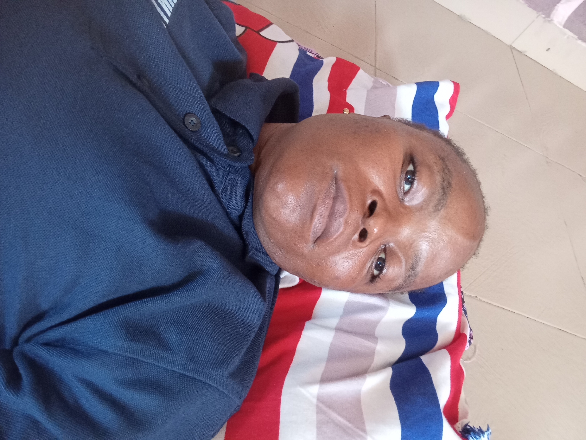 A person is lying down, their head resting on a striped pillow. They are wearing a dark blue collared shirt with a button closure. The pillow has alternating red, white, and blue stripes. The background appears to be a tiled floor. The image is taken from a high angle, looking down at the person. The person's face is partially visible, with their eyes open. The scene is indoors, likely in a residential setting. The location is Kason, Nigeria.