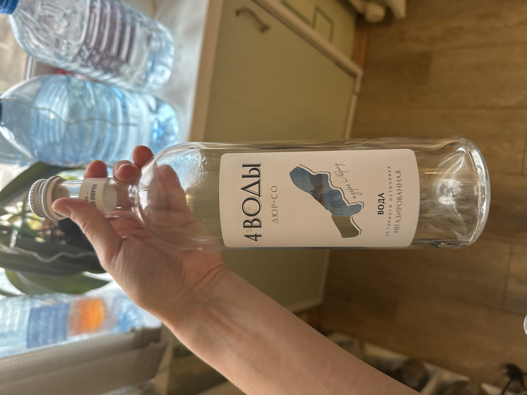 The image depicts a hand holding a glass bottle of "4 VODY" water, originating from a mountain spring near Lake Abrau. The bottle is labeled "ДЮР-СО" and indicates that the water is non-carbonated. The setting appears to be indoors, with other water bottles visible in the background. The city of Kladovitsy, Russia is not directly visible or identifiable in the image.