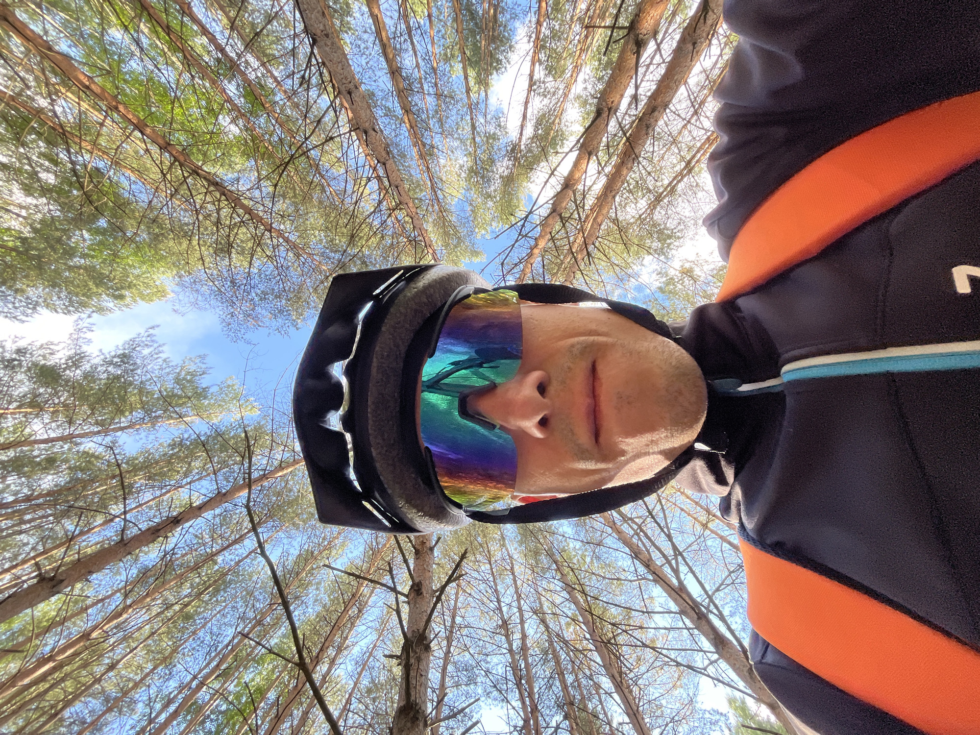 A low-angle selfie captures a person looking up into a dense pine forest canopy under a clear, bright blue sky, likely in Chaikovsky, Russia. The individual, appearing male, is in the foreground, wearing a dark cycling helmet and large, multi-colored reflective sports sunglasses. Only the lower part of their face—nose, mouth, and chin—is visible, framed by the helmet and glasses. The sunglasses reflect a distorted view of the surrounding trees and sky.
The person is dressed in a dark blue long-sleeved athletic top, which features a prominent orange diagonal strap across the chest or shoulder, possibly part of a backpack or harness. A thin light blue and white stripe is visible at the collar area. On the right side of the top, a partial white logo resembling an "N" can be seen.
The background is dominated by numerous tall, slender pine trees, their trunks and branches extending upwards, creating a natural pattern against the sunny sky. The foliage is a mix of green and brown, with sunlight filtering through, suggesting a clear day, either morning or afternoon. The overall scene conveys an immersive outdoor experience, likely during an activity such as cycling or mountain biking, in a tranquil natural environment.