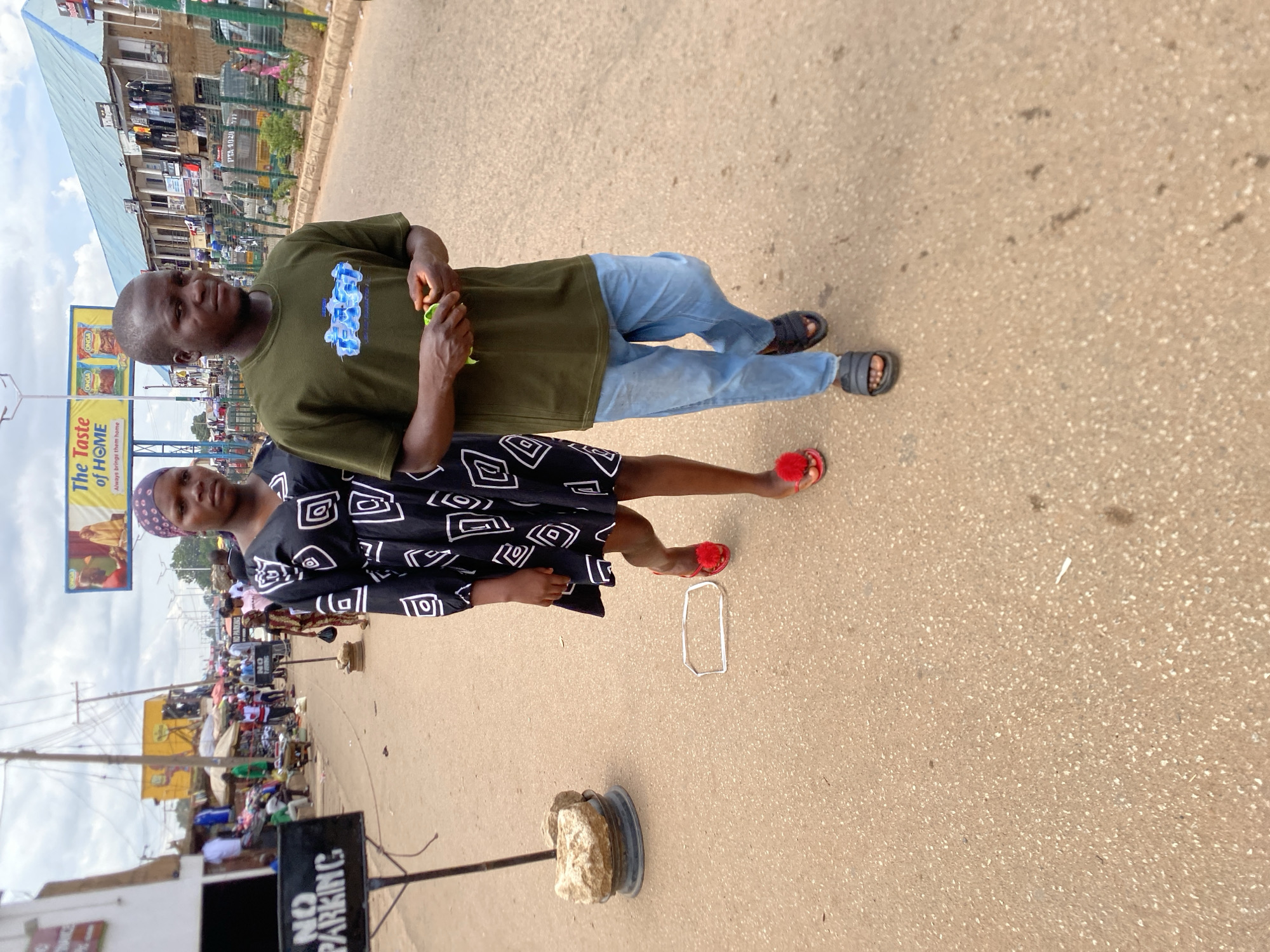 This image captures a street scene in Bukuru, Nigeria, under an overcast sky. In the foreground, two dark-skinned individuals walk towards the viewer on a light tan, paved road. On the left, a woman wears a black dress with white geometric patterns, a patterned headscarf (purple/blue and white dots), and red open-toed sandals with red spherical embellishments. Her right foot is slightly elevated mid-stride. On the right, a man wears a dark olive green t-shirt with a light blue stylized graphic, light blue baggy jeans, and dark grey slide sandals. He holds a crumpled light green object in his hands. Both individuals maintain neutral expressions, looking generally forward.
To the left of the woman, a "NO PARKING" sign with black text on a white panel is mounted on a dark pole, stabilized by stacked rocks and a metal ring base. Near the woman's feet, a white, rectangular, hollow object lies flat on the road.
The midground and background reveal a bustling street environment. On the left side of the road, various market stalls, umbrellas, and numerous people are visible in front of buildings, including a white structure and a prominent yellow building. Utility poles and overhead wires crisscross the sky. Another smaller "NO PARKING" sign is partially visible further down this side of the street. On the right side, a long, two-story building with a light blue corrugated metal roof features multiple shopfronts displaying clothing. A low concrete curb with small green plants runs along this side of the road. Further down the street, more people and vehicles, including a yellow tricycle (keke napep), are present.
An overhead billboard spans the street, displaying "The Taste of HOME" and "Always brings them home," featuring images of food products, specifically "ONGA" brand packets. The overall lighting is diffused daylight.