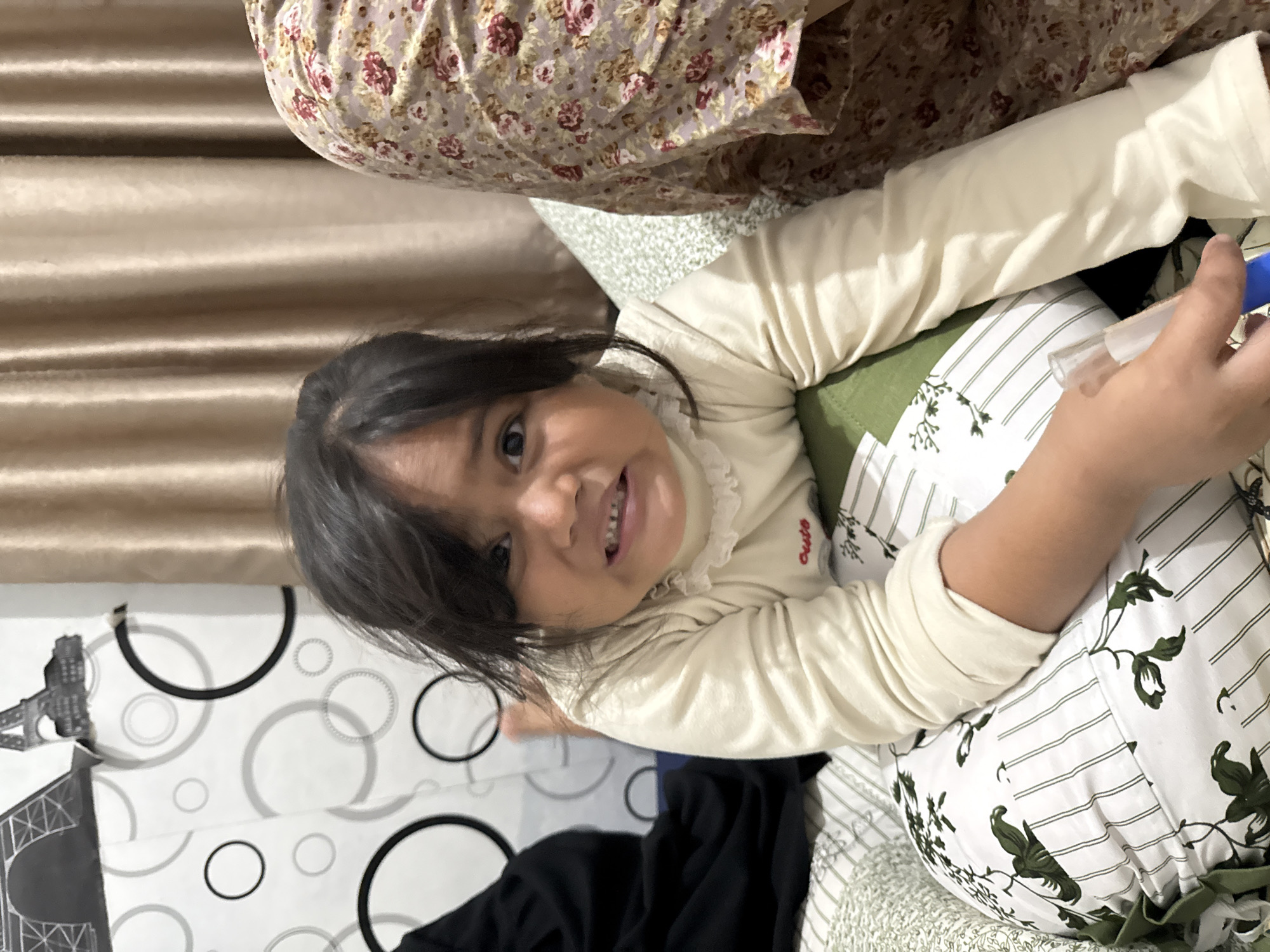 A young child with dark hair is lying on their back, looking up at the camera with a wide smile. The child is wearing a long-sleeved cream-colored shirt with a ruffled neckline. They are nestled on a bed with white and green striped bedding featuring a floral pattern. A hand holding a blue object, possibly a toy or utensil, is visible near the child's arm. In the background, a curtain with horizontal folds can be seen, and part of another person wearing a floral-patterned garment is also visible. The overall impression is a casual, indoor setting, likely a home, possibly in Babahjurong, Indonesia. The child's expression conveys happiness and engagement.