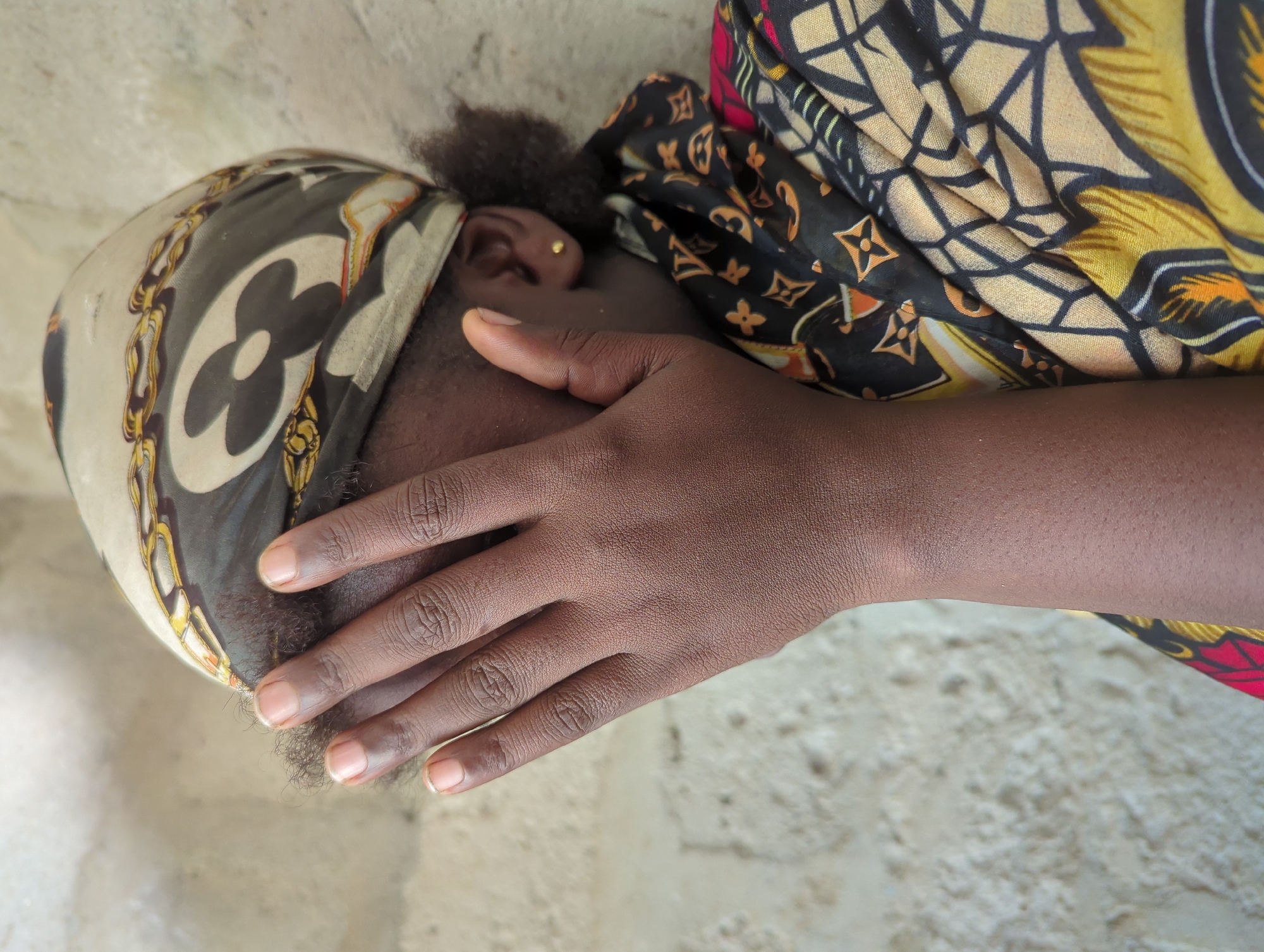 A close-up photograph depicts a person of African descent with dark skin and dark hair, partially obscured by a headscarf and hand. The individual is wearing a patterned headscarf featuring a repeating floral or clover-like design and a gold chain pattern, positioned over the left side of their head. The person's hand covers the lower half of their face. The ear is visible and adorned with a small gold stud earring. The person is wearing a colorful patterned garment with black, yellow, and red elements. The background is a textured, neutral-colored surface, possibly a wall. The location is Elele Alimini, Nigeria.