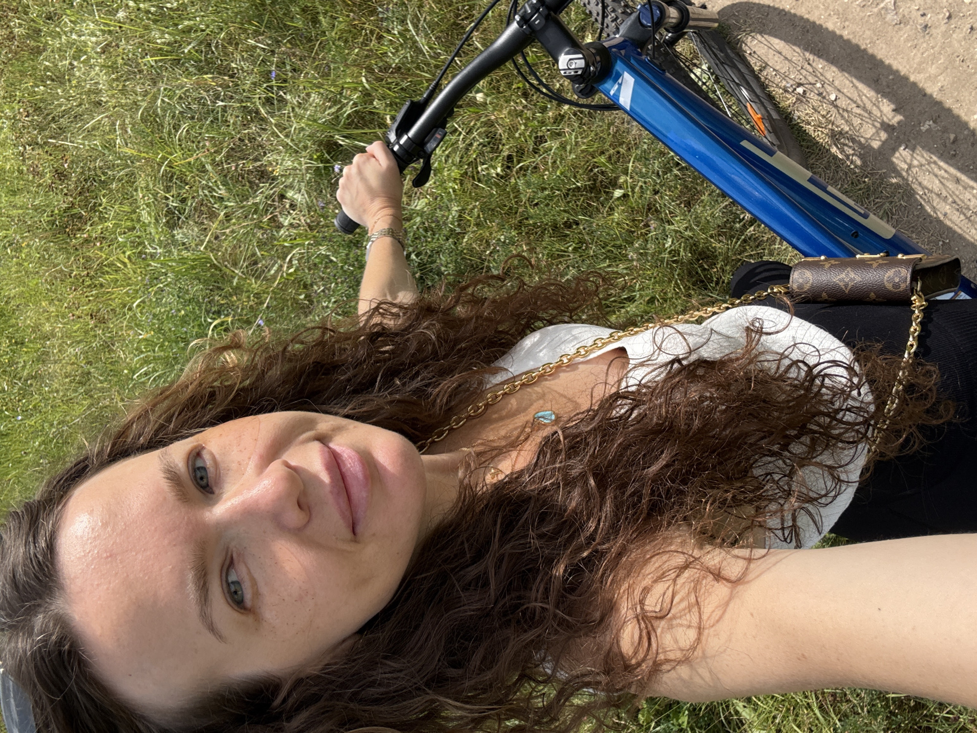 A woman with curly brown hair is taking a selfie while sitting on a blue bicycle outdoors. She is wearing a white tank top and a gold chain necklace. The background is a grassy area with some dirt path. The image does not contain any information that can determine the location to be Moscow, Russia.