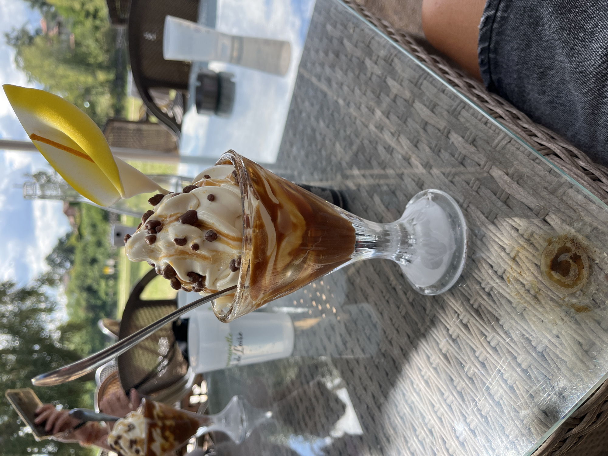 The image depicts an outdoor dining scene in Saint-Just-Saint-Rambert, France. In the foreground, a glass of ice cream with caramel sauce and chocolate shavings is prominently featured on a glass-topped table with a woven base. A silver spoon rests in the ice cream. To the left, another glass of ice cream is partially visible, along with a white cup. In the background, a person holds a smartphone, and other patrons are seated at tables. The environment suggests a sunny day with trees and buildings visible in the distance. The overall atmosphere is casual and leisurely, typical of an outdoor cafe or restaurant setting.