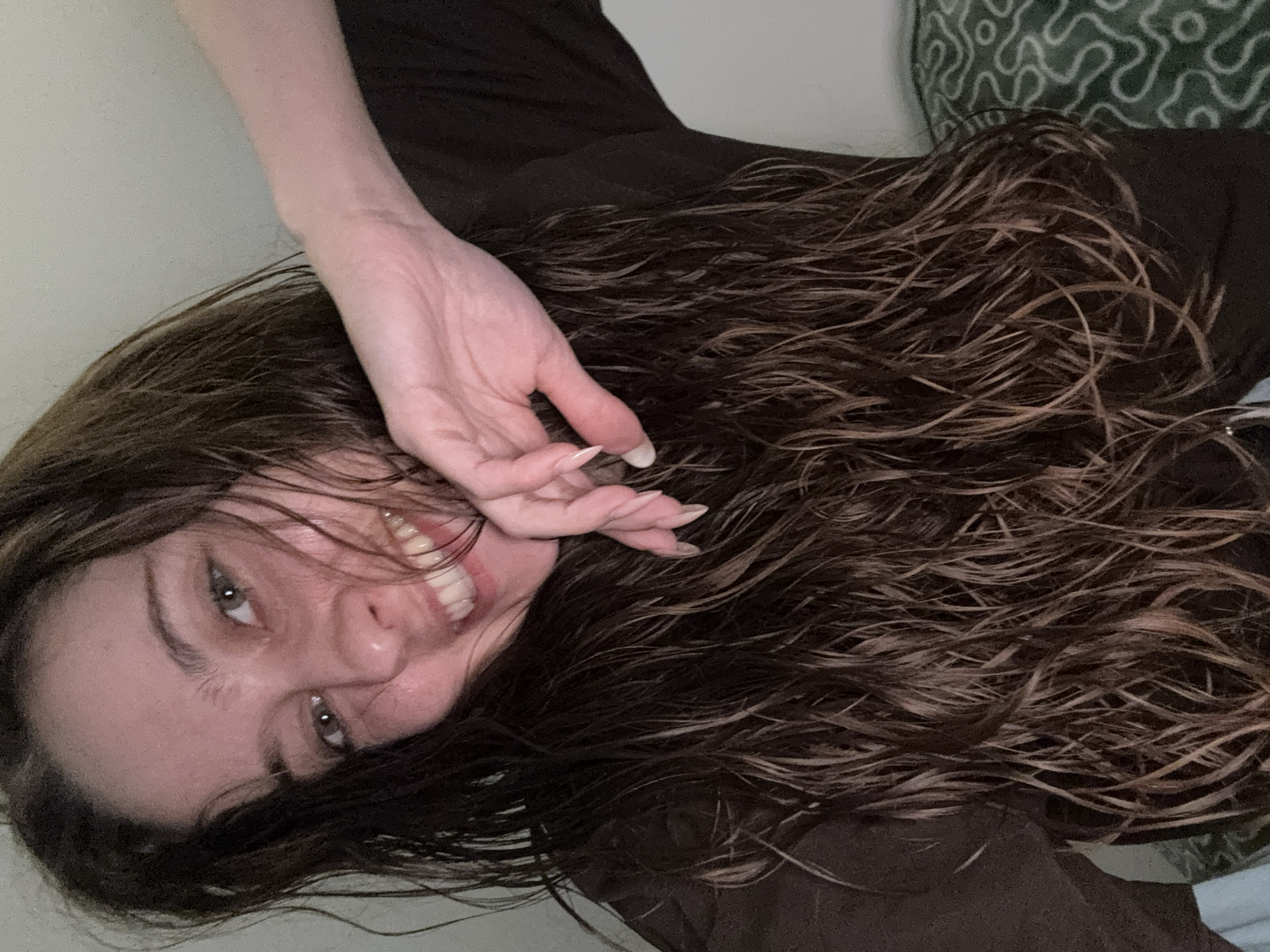 A woman with long, dark, wet hair is lying down, facing the camera. Her hand is near her face. She is smiling, showing her teeth. She is wearing a dark top. The background includes a patterned pillow and a wall. The location is unknown and cannot be determined to be Ramat Gan, Israel.
