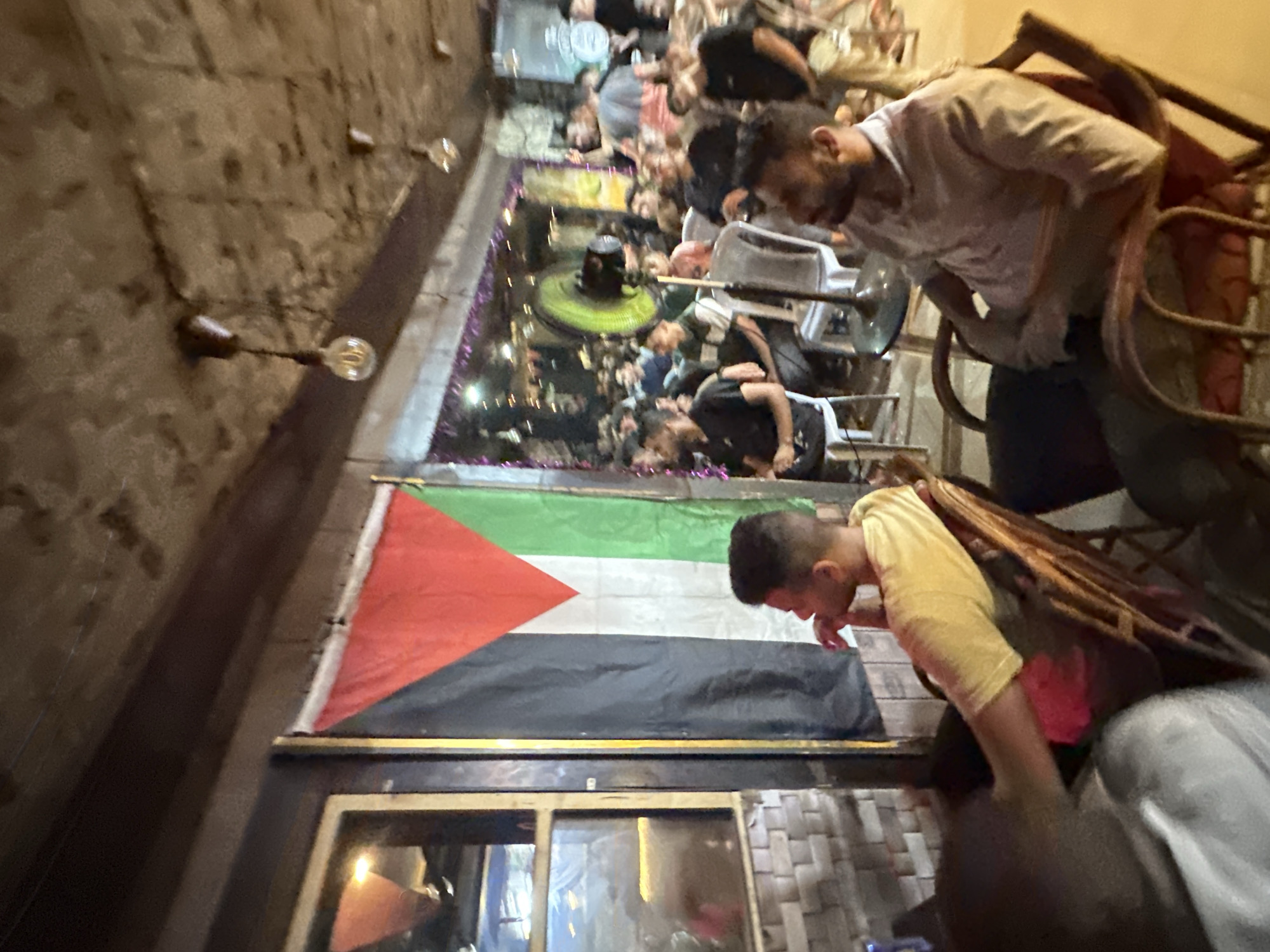 In the city of Gaza, Palestinian Territories, a dimly lit, indoor space with a rough, concrete ceiling features several exposed light bulbs hanging from wires. A Palestinian flag is prominently displayed on the left side of the frame, hanging vertically against a wall adorned with brick-like tiles. In the foreground, two men are seated in chairs. The man on the left, wearing a yellow t-shirt, is looking to his left, while the man on the right, in a light-colored collared shirt, is facing forward, looking down and to his right. In the background, a crowd of people, predominantly men, are seated, facing what appears to be a screen or stage, with a large green fan visible in the center. The overall atmosphere suggests a gathering or viewing event in a casual setting.