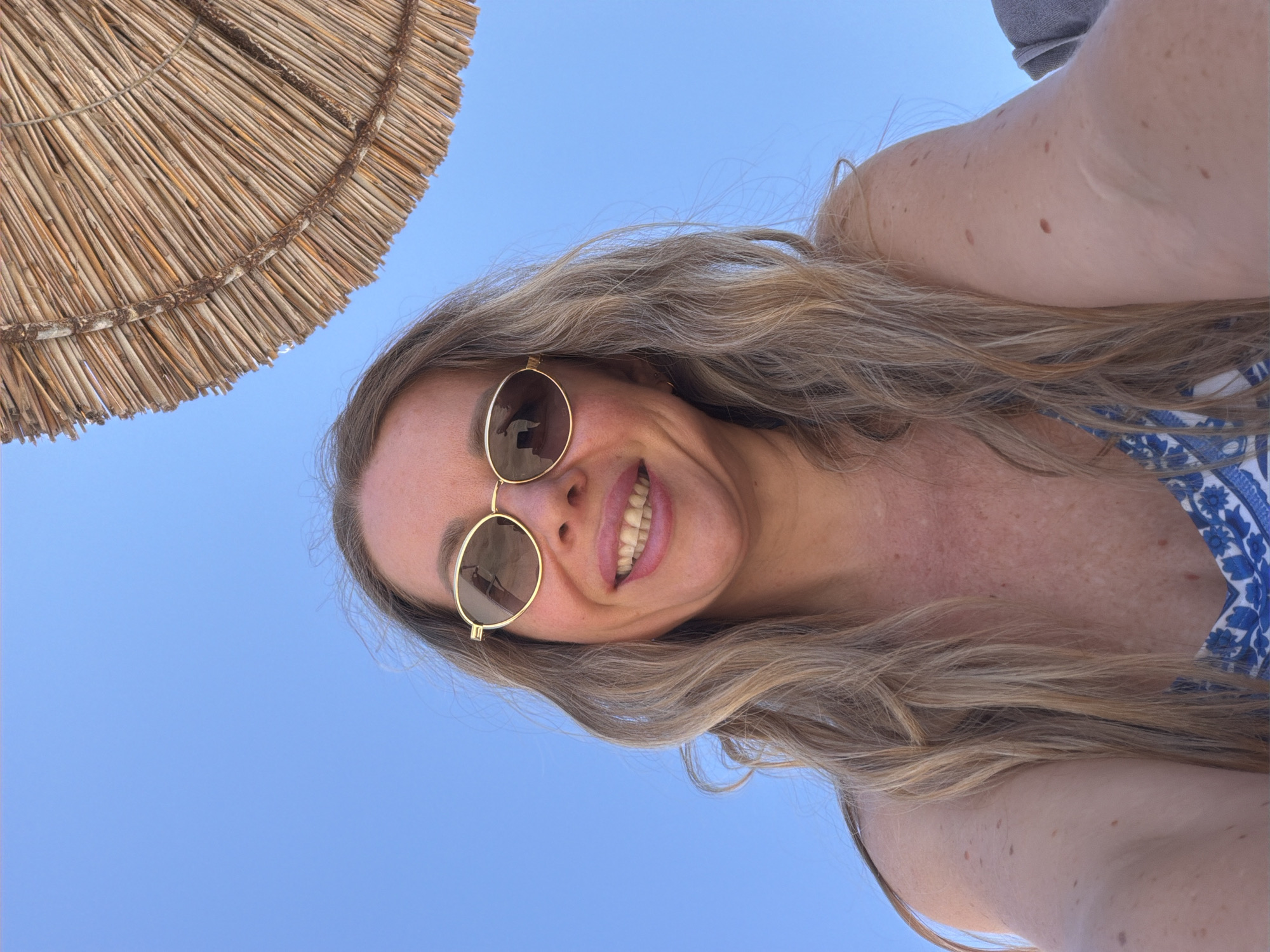 A woman smiles at the camera from below, under a thatched umbrella against a clear blue sky. She has blonde hair, is wearing sunglasses, and is dressed in a blue and white patterned swimsuit. The shot appears to be taken outdoors, likely in a sunny, tropical location.