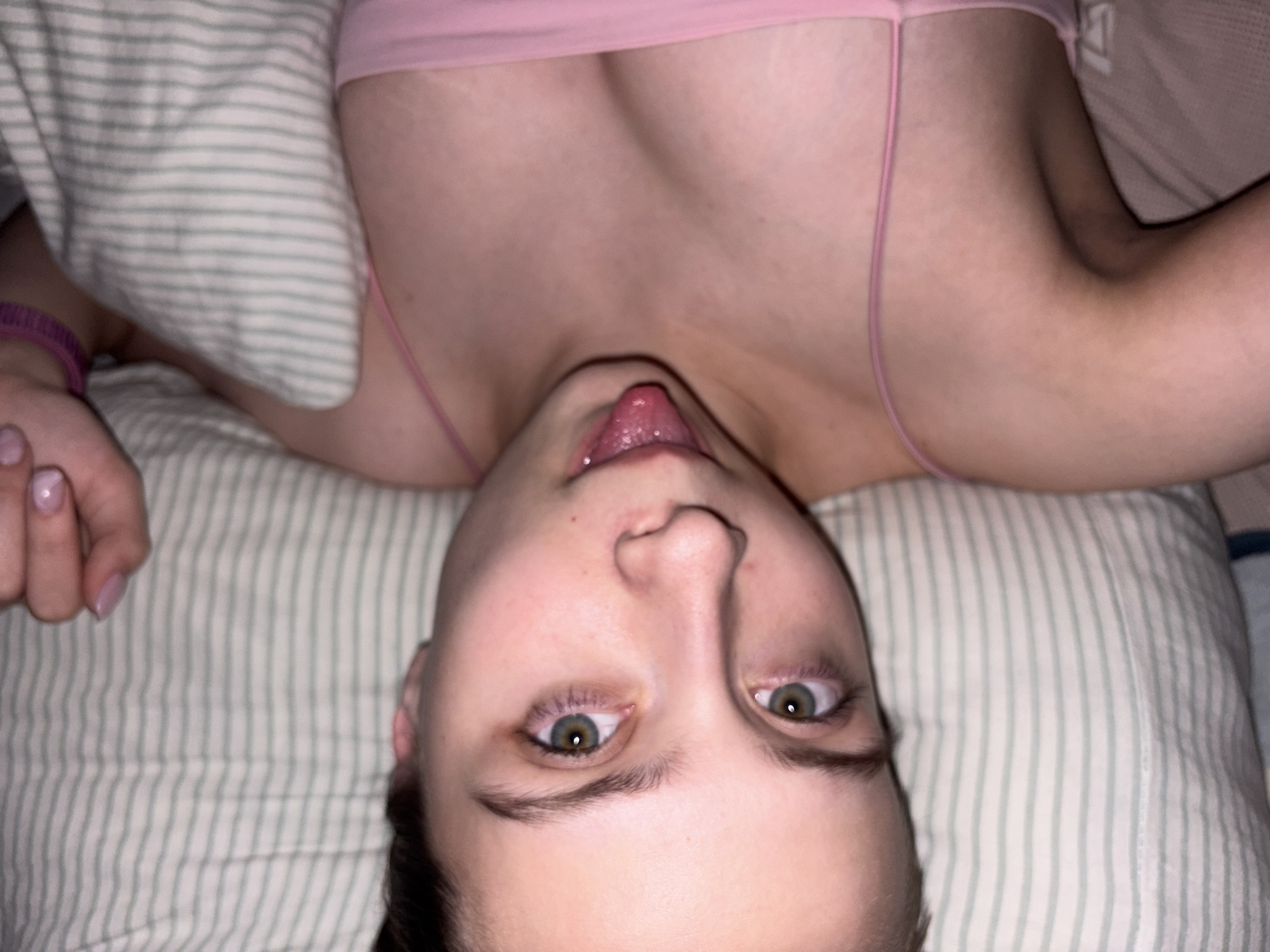 An inverted close-up shot captures a person lying down, seemingly on a bed or pillow with green and white striped bedding. The subject, likely female, is positioned with their head at the bottom of the frame, looking up towards the camera. Their tongue is slightly extended, and their green eyes are wide open, appearing somewhat red around the whites.
The person is wearing a pink spaghetti strap top or bra, part of which is visible across their chest and shoulders. A purple wristband is worn on the left wrist, and their fingernails are painted a light pink. The overall posture suggests a playful or relaxed moment. The lighting is soft, indicating an indoor setting, possibly a bedroom. No visible text is present in the image. The scene takes place in Прикамский, Russia.