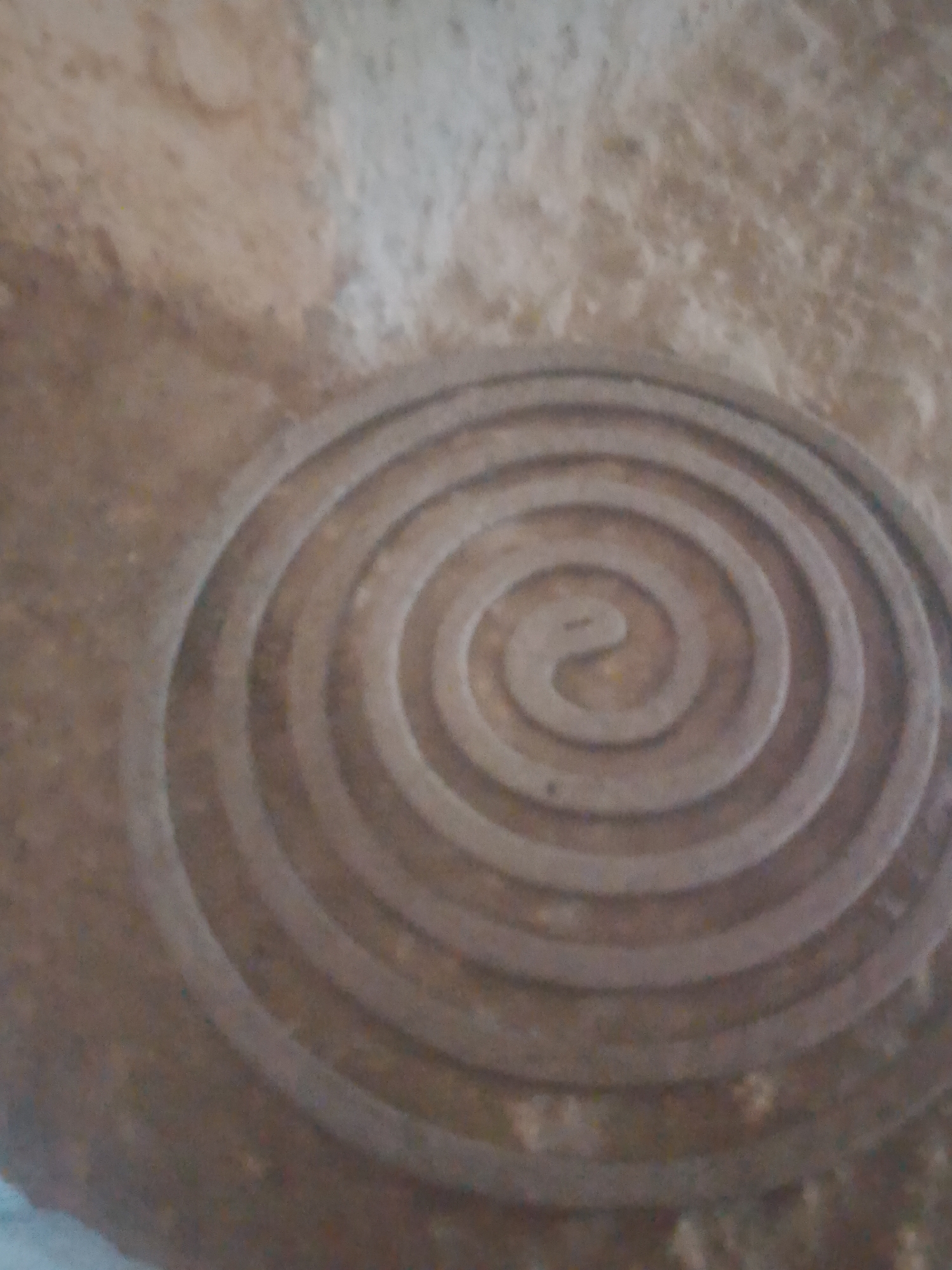 The image shows a close-up view of a spiral carving in a textured, stone-like surface. The spiral is concentric and appears to be etched or carved into the material. The surface has a rough, uneven texture with variations in color, ranging from light beige to darker brown. Some small, irregularly shaped particles are visible embedded in the surface. The lighting suggests an indoor or shaded outdoor environment, casting subtle shadows that emphasize the depth of the carving. The carving is the central and dominant subject of the image. There are no people, actions, or interactions depicted. The location is identified as Jauro Yinu, Nigeria.