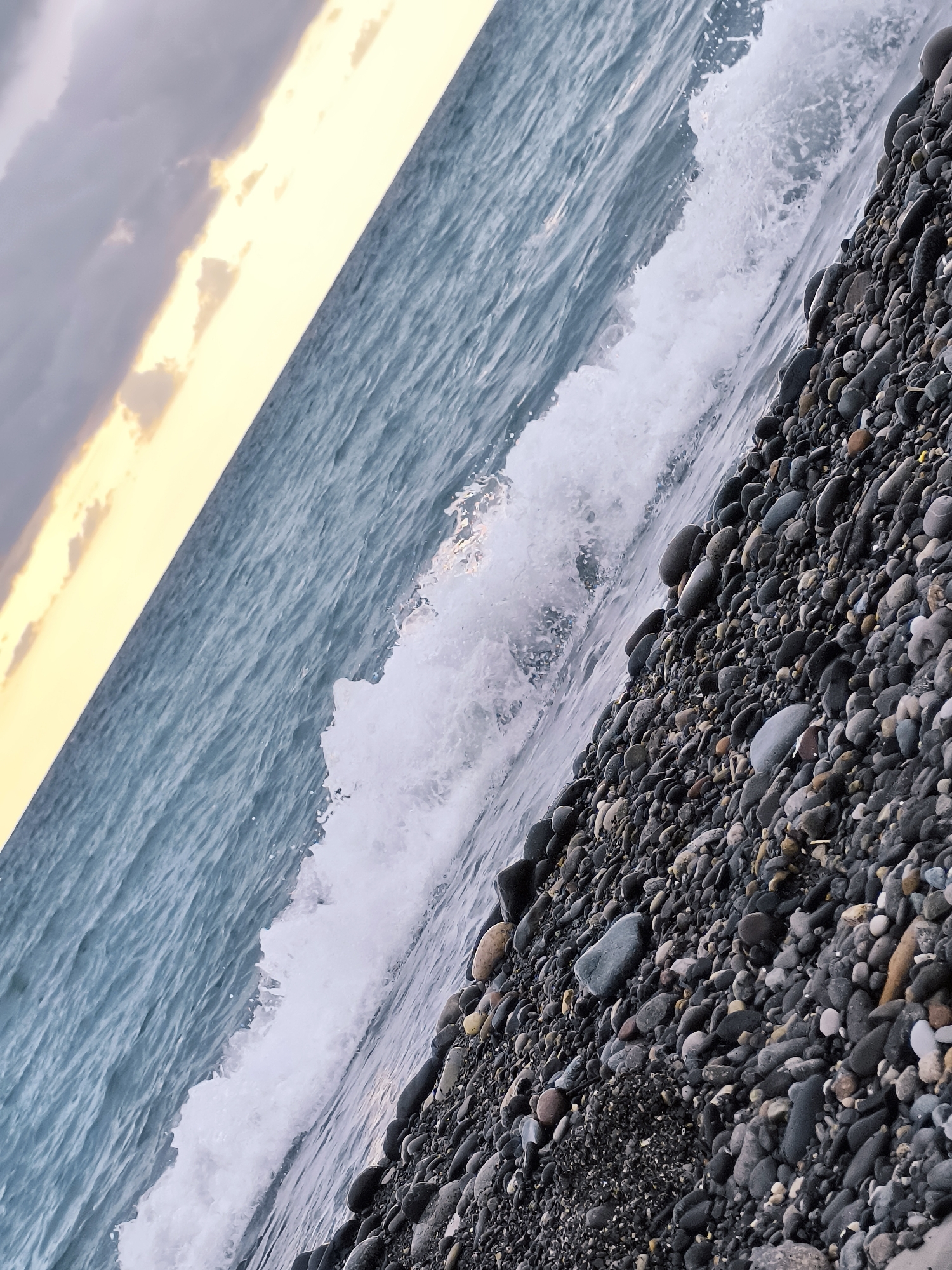 The image depicts a slightly tilted view of a pebble beach meeting the sea. The water is a muted blue-gray, with a breaking wave creating white foam near the shore. The beach is covered in numerous smooth, dark pebbles of varying sizes. In the upper left of the frame, the sky is a mix of grey clouds and a soft yellow band of light, suggesting either dawn or dusk. The location is identified as Zemo Kvariati, Georgia. There are no people or animals visible in the scene. The environment is a natural coastal landscape.