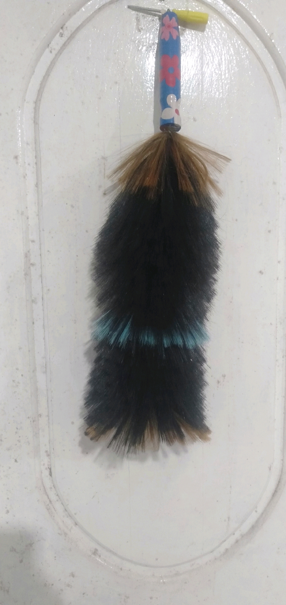 In the city of Cikoneng, Indonesia, a decorative dust brush hangs on a white, embossed door. The brush has a blue handle with hand-painted pink and white flowers. A yellow object, possibly a pen cap, is attached to the top of the handle. The brush itself is made of black bristles with a band of teal bristles in the middle and brown tips. The door appears to be an interior door, possibly in a home. The lighting is dim, suggesting it may be indoors.