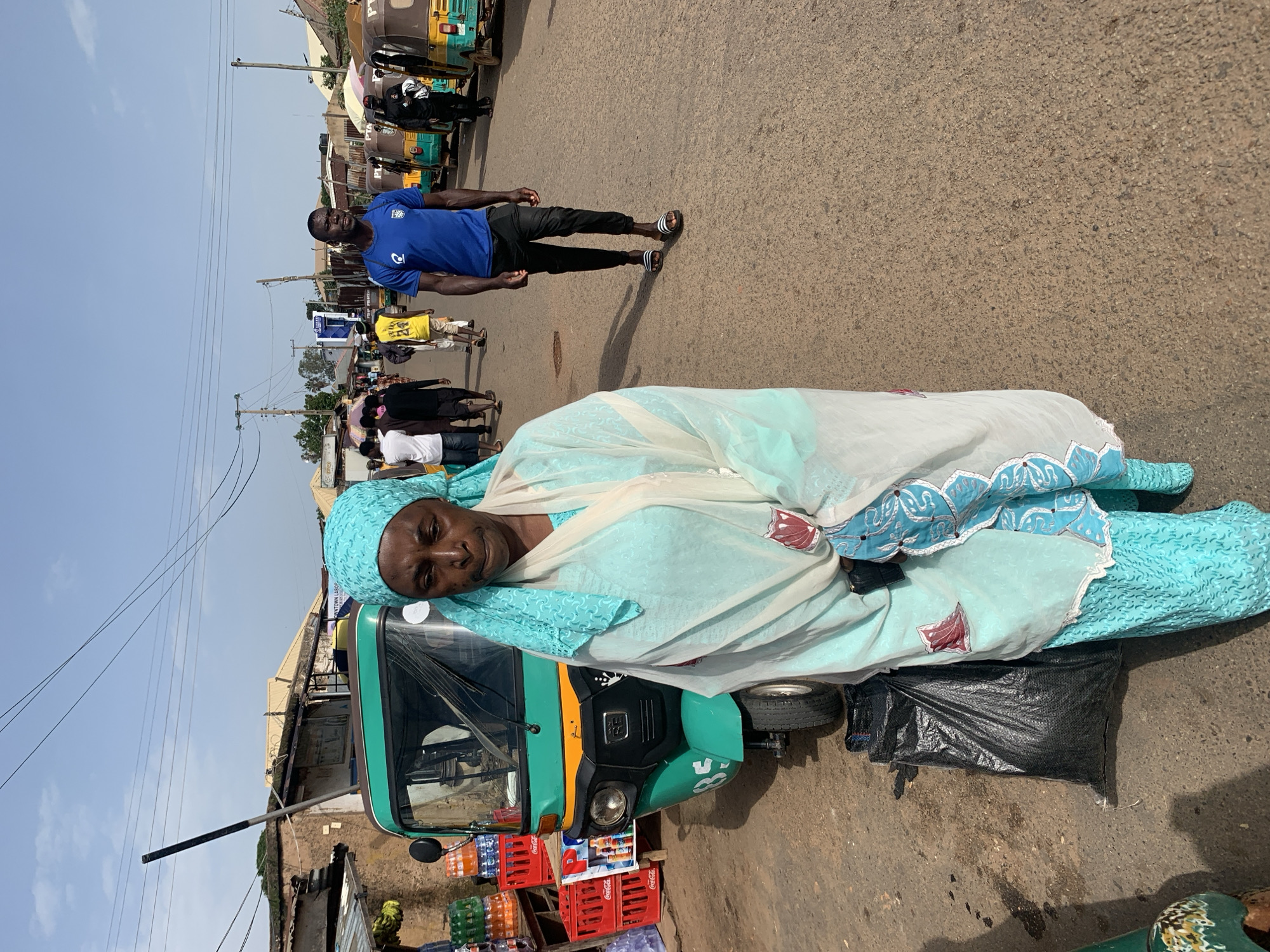 In Gura Top, Nigeria, a woman in a light blue headscarf and matching patterned dress stands near a teal and yellow motorized tricycle. Her white and blue patterned shawl is draped over her shoulders. In the background, a man in a blue t-shirt and black pants walks away from the camera, while other pedestrians and more tricycles line a dusty road. Buildings with corrugated metal roofs are visible on either side of the street, suggesting a marketplace or bustling urban area. The sky is clear and blue, with power lines stretching across the frame. A black plastic bag sits on the ground next to the woman and the tricycle.