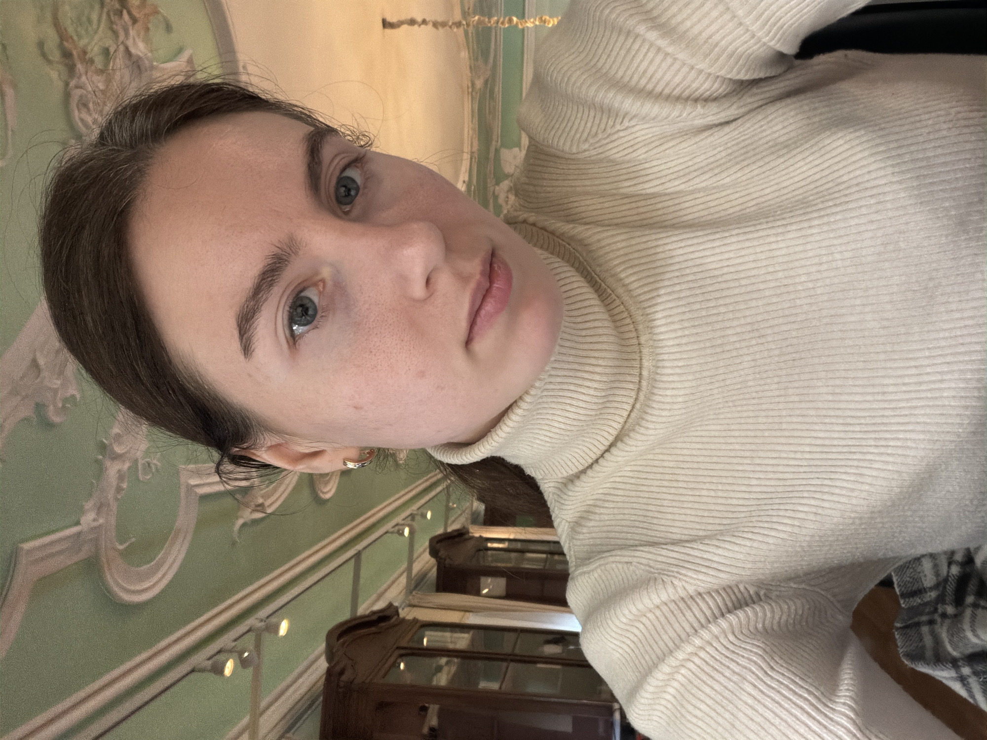 A young woman with fair skin, brown hair, and blue eyes is the primary subject of the photograph. She is wearing a ribbed turtleneck sweater. The background indicates she is indoors, possibly in a historic building in the city of Saint Petersburg, Russia. There is intricate baroque-style wall ornamentation, light green in color, a common feature in historical buildings in Saint Petersburg. Beneath are display cases, possibly indicating she is in a museum or a historical site open to the public.