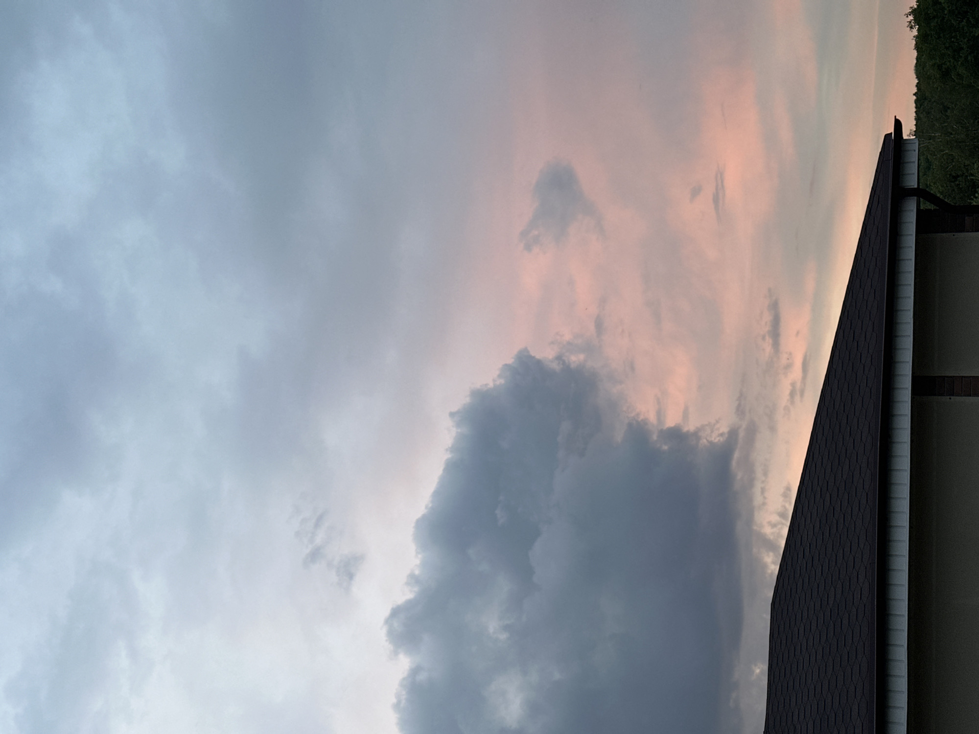The image shows a sky filled with clouds, varying in color from light blue-gray to darker gray and pinkish hues. A dark-colored rooftop with a repeating pattern of shingles occupies the right side of the frame. Adjacent to the roof is a light-colored building exterior. Green foliage is visible in the upper right corner. The scene appears to be taken outdoors, likely at dusk or dawn based on the lighting. There is no verifiable data to suggest the city of Nizhny Novgorod, Russia, is the location.