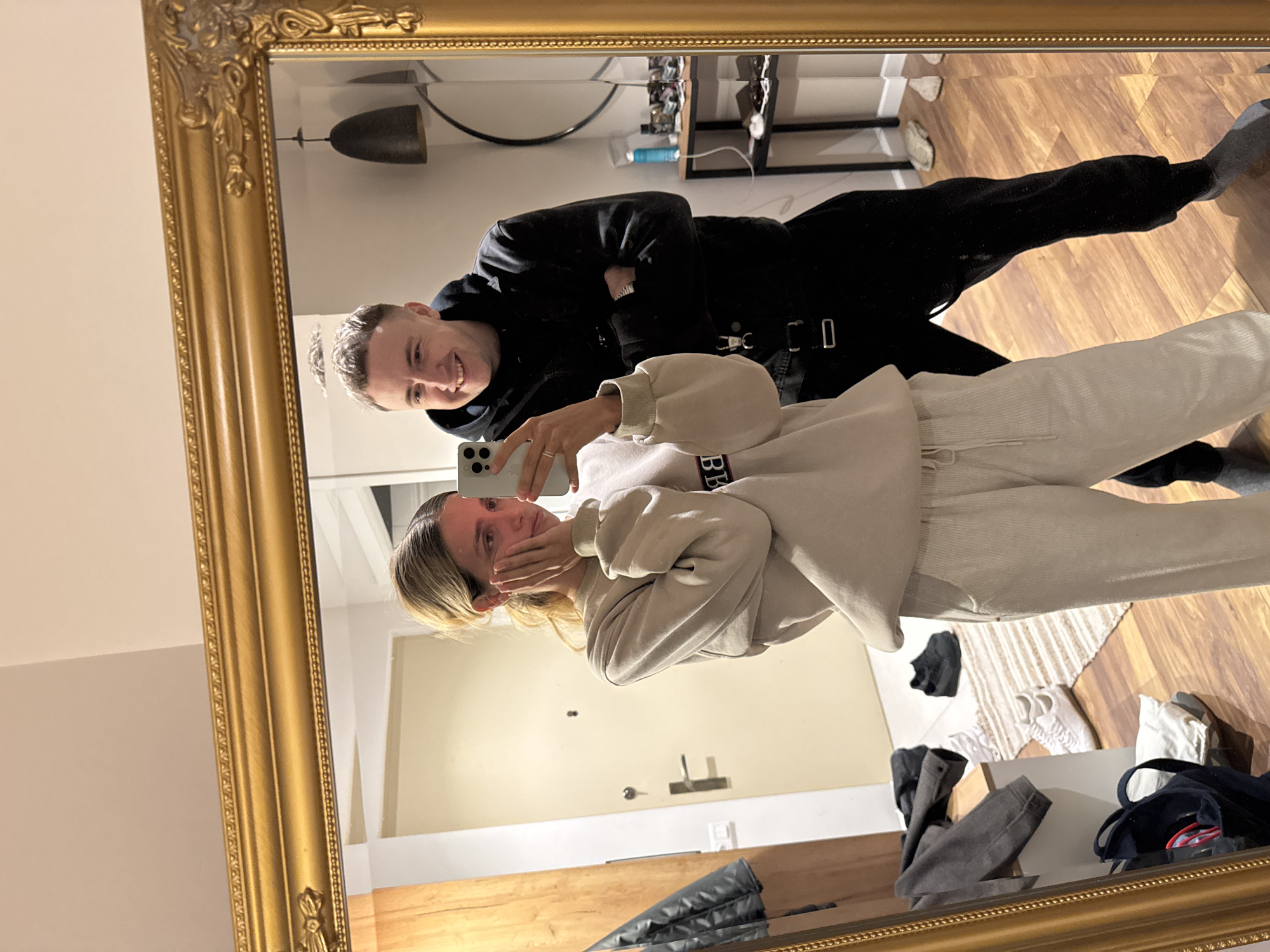 A framed mirror reflects two people taking a selfie in what appears to be a residential setting. The mirror's ornate gold frame is partially visible. A woman is holding a smartphone up to capture the image, partially obscuring her face with her hand. She is wearing a light beige sweatshirt and matching pants. Behind her, a man wearing a black hooded sweatshirt and pants is visible. He is smiling at the camera with his arms crossed.
The reflection shows a wooden floor, a door in the background, and various items scattered on the floor near the base of the mirror, including clothing and shoes. A shelf with miscellaneous objects is visible in the background. A ceiling light fixture is also reflected in the mirror. The location is unknown.