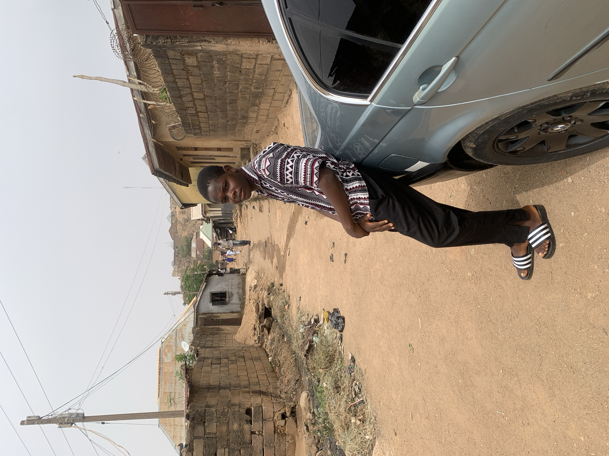 A young man in a patterned shirt and black pants poses next to a light blue car on a dirt road. He wears striped sandals. The car is parked near a brick building topped with barbed wire. Behind the car and the man, a street scene unfolds with other buildings, some with corrugated iron roofs. There's scattered trash and debris alongside the road. In the background, a townscape extends toward a hazy sky. The image is taken in Jos, Nigeria.