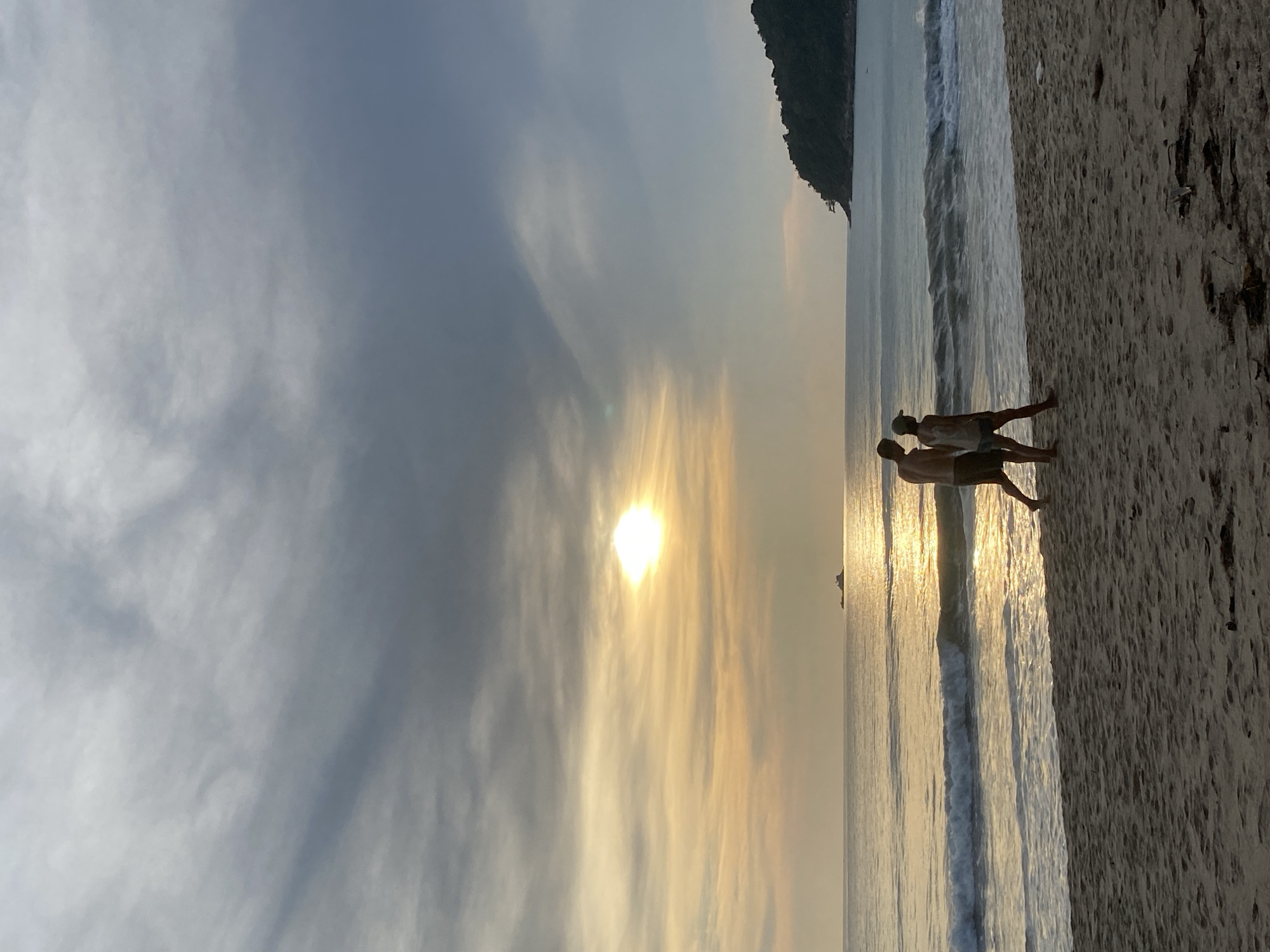 The image depicts a sunrise on Bang Tao Beach in Choeng Thale, Thailand. Two figures are standing near the water's edge. The sky is a blend of gray and golden hues, with the sun partially obscured by clouds. A landmass is visible in the distance. The foreground is sandy beach with visible details in the sand. Waves are gently breaking on the shoreline. The image is oriented vertically.