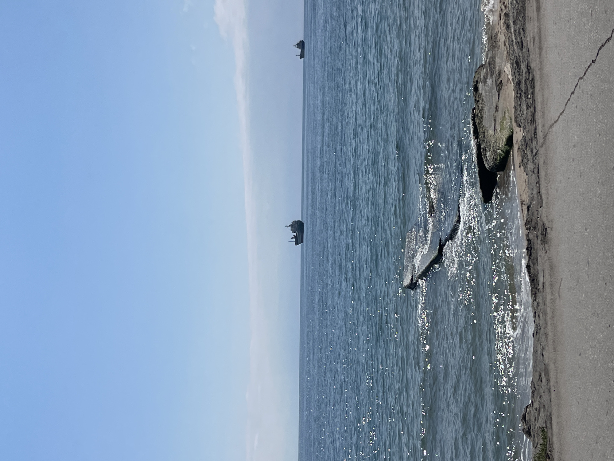 The photograph depicts a seascape in Varna, Bulgaria, featuring a body of water, likely the Black Sea, occupying most of the frame. The sky, a light blue hue, is visible on the left side. Two vessels are seen on the water. The right side shows a shoreline with concrete or stone structures and vegetation. The perspective is from near the shoreline, giving a sense of being at the edge of the water. Sunlight reflects off the water's surface.