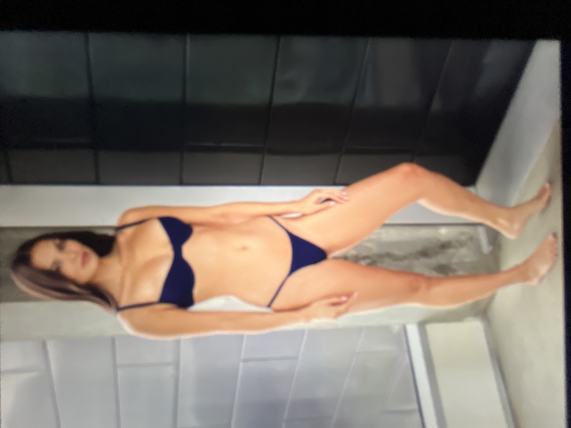 The image shows a full-body shot of a woman wearing a dark blue bikini. She is standing in what appears to be a modern bathroom or changing room, with tiled walls and a dark, textured wall on the right. The woman has long, dark hair and is looking towards the camera. Her pose is relaxed, with her weight shifted to one leg. The lighting is soft, creating a slightly diffused effect on the image. The background is out of focus, with the focus primarily on the woman. There is no visible indication of the location being in Sofia, Bulgaria.