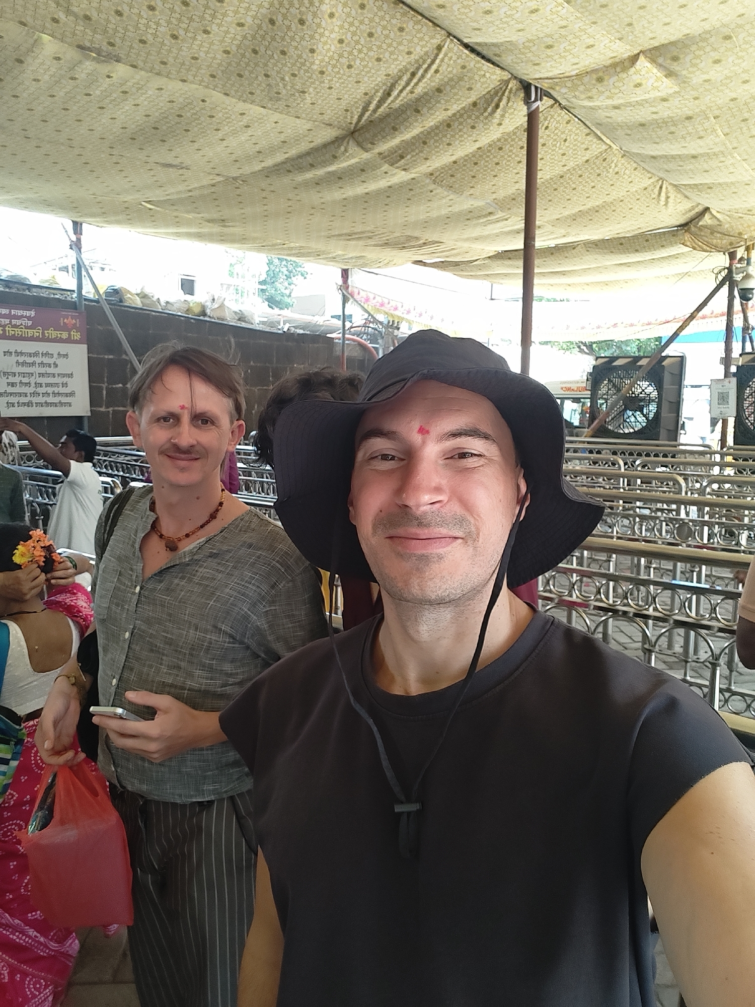 The image depicts a selfie-style photograph taken in a covered public area, likely a temple complex or a crowded civic space in Kolhapur, India.
The primary subject in the foreground right is a male with light skin, short dark hair, and a slight stubble. He wears a black bucket hat and a black, short-sleeved crew-neck t-shirt. A black cord with a toggle hangs from his neck. A red tilak mark is present on his forehead, and he smiles directly at the camera.
Positioned slightly behind and to the left is another male, with light skin, light brown hair, a mustache, and a red tilak on his forehead. He wears a light grey, long-sleeved button-up shirt with rolled-up sleeves, dark grey pinstriped trousers, and a brown beaded necklace. He holds a silver smartphone in his left hand and a small red plastic bag in his right, also smiling at the camera.
In the mid-ground left, a partially visible female individual is seen wearing a white top and a pink patterned garment, holding orange and yellow flowers, typically used as offerings. Further left and behind her, a male in a white garment is partially visible, moving towards the left.
The environment features an overhead fabric canopy, light-colored with a repeating circular pattern, supported by brown poles. In the background, silver-colored metal crowd control barriers are visible. A dark, possibly stone, wall forms the backdrop, upon which a white sign with black Marathi text is affixed. The legible text includes "श्रीमंत छत्रपती शाहू महाराज," referencing a prominent historical figure from Kolhapur. Two large, dark industrial fans are mounted above the crowd barriers on the right, alongside a security camera attached to a pole. Green foliage is discernible in the far background, suggesting an open-air area beyond the immediate structure.