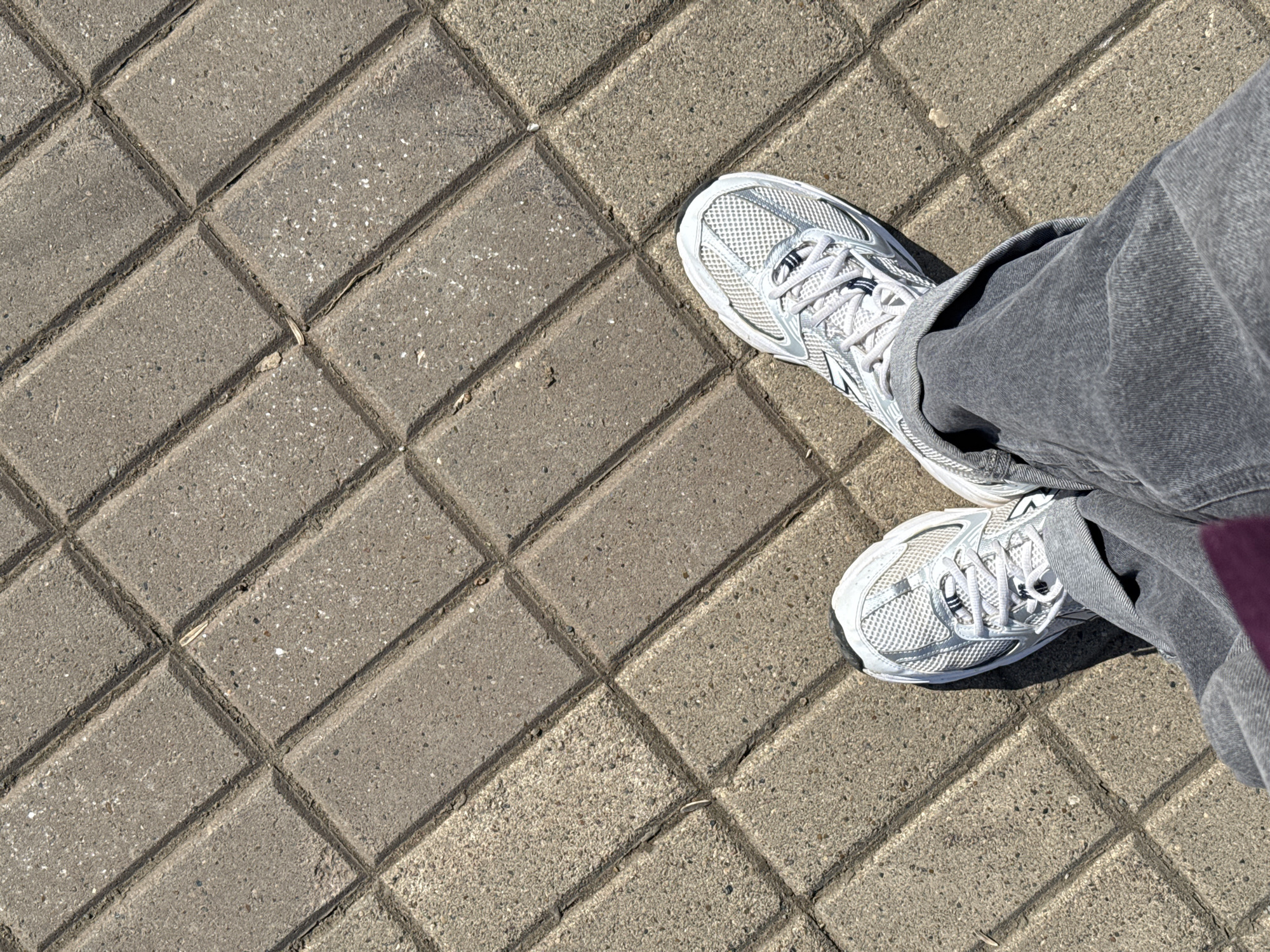 The image shows a first-person perspective of a person standing on a paved surface. The surface is composed of rectangular concrete bricks laid in a diagonal pattern. The bricks are gray with darker grooves outlining them. The person is wearing gray jeans and white sneakers with white laces. The location is undetermined, but the prompt suggests it is in Izhevsk, Russia. There are no other people, subjects, objects, actions, or interactions visible in the image.