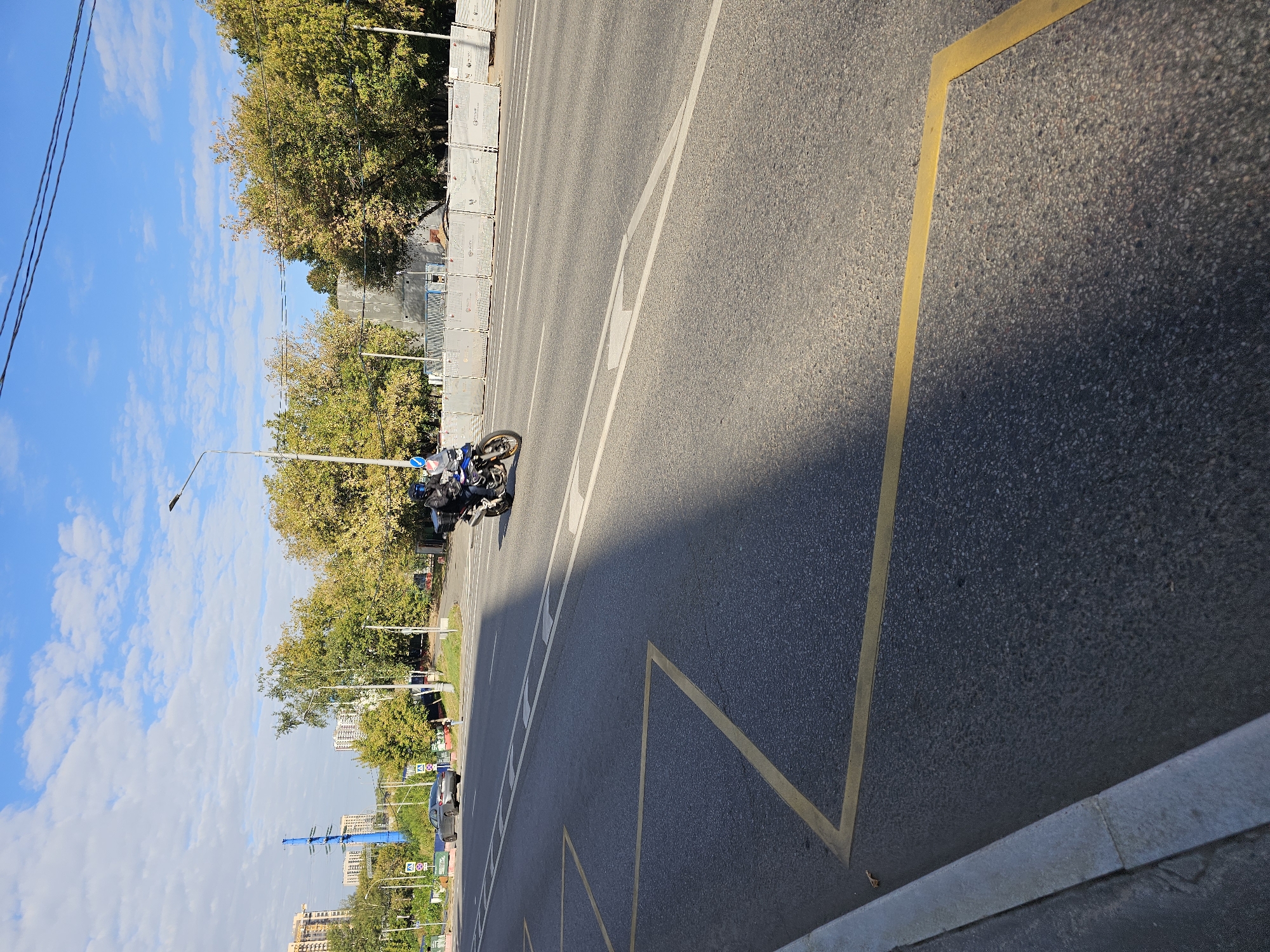 A motorcycle rider in dark clothing and a blue helmet navigates a road in Moscow, Russia. The rider is on a dark-colored motorcycle with a black storage box on the back, moving from left to right in the frame. The road is asphalt with clear yellow lane markings, including diagonal lines forming a junction. A gray car is visible on the opposite side of the road, heading towards the motorcycle. In the background, there are trees with autumnal foliage, buildings, and a tall blue power transmission tower. The sky is blue with scattered white clouds. A shadow from an unseen object falls diagonally across the road. The scene appears to be a street in a city, possibly near a construction site indicated by temporary fencing.