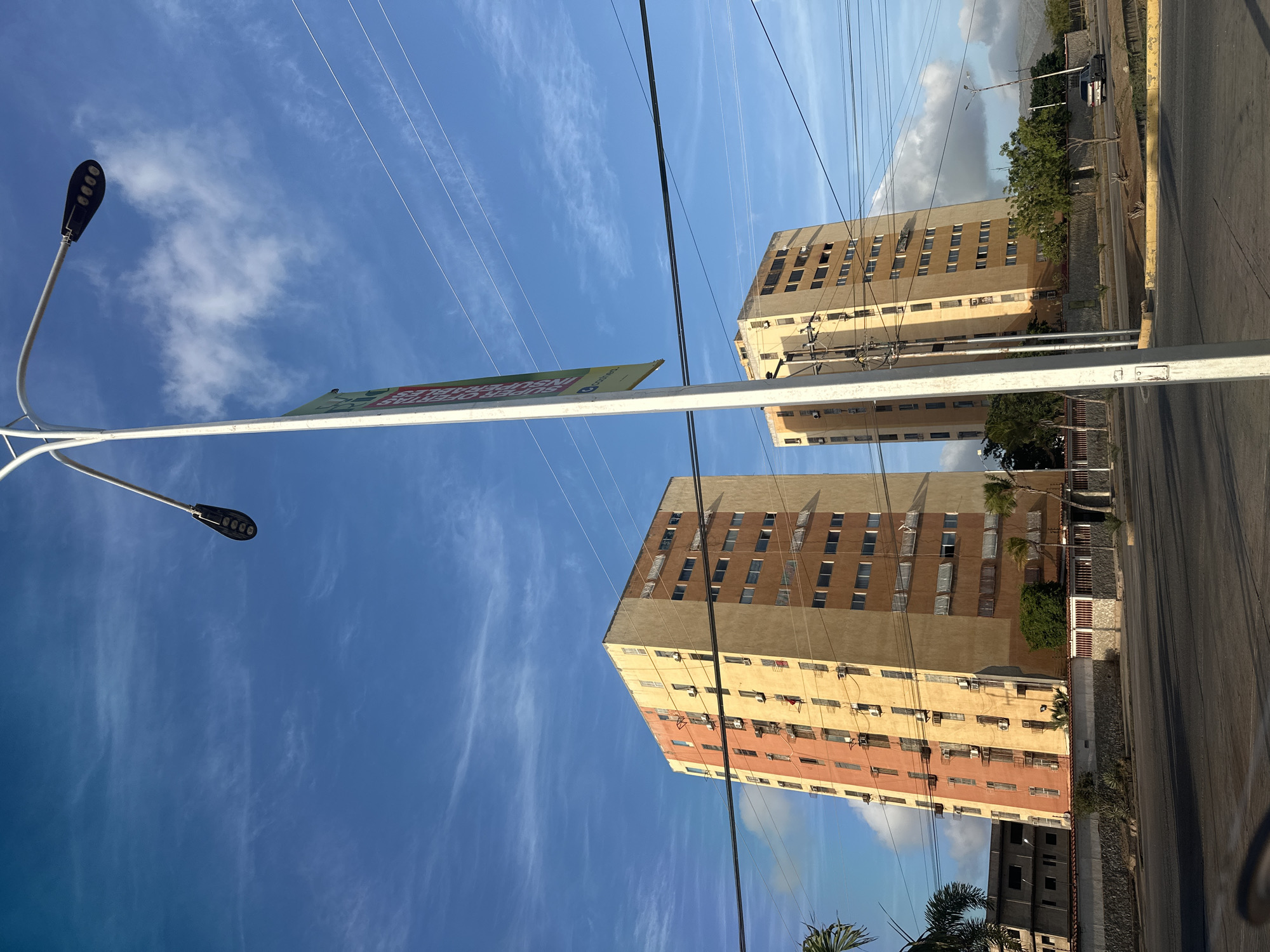 This image shows two tall, multi-story apartment buildings in Juan Griego, Venezuela, under a bright blue sky with wispy white clouds. A tall lamppost with a banner is positioned in the foreground, slightly off-center. The buildings are light brown and orange, with numerous windows visible on their facades. Power lines crisscross the sky above the buildings. The street in front of the buildings appears to be a road with some traffic. Palm trees and other vegetation are scattered around the base of the buildings and along the roadside. In the background, hills or mountains are visible in the distance. The overall scene depicts an urban environment in daylight.
