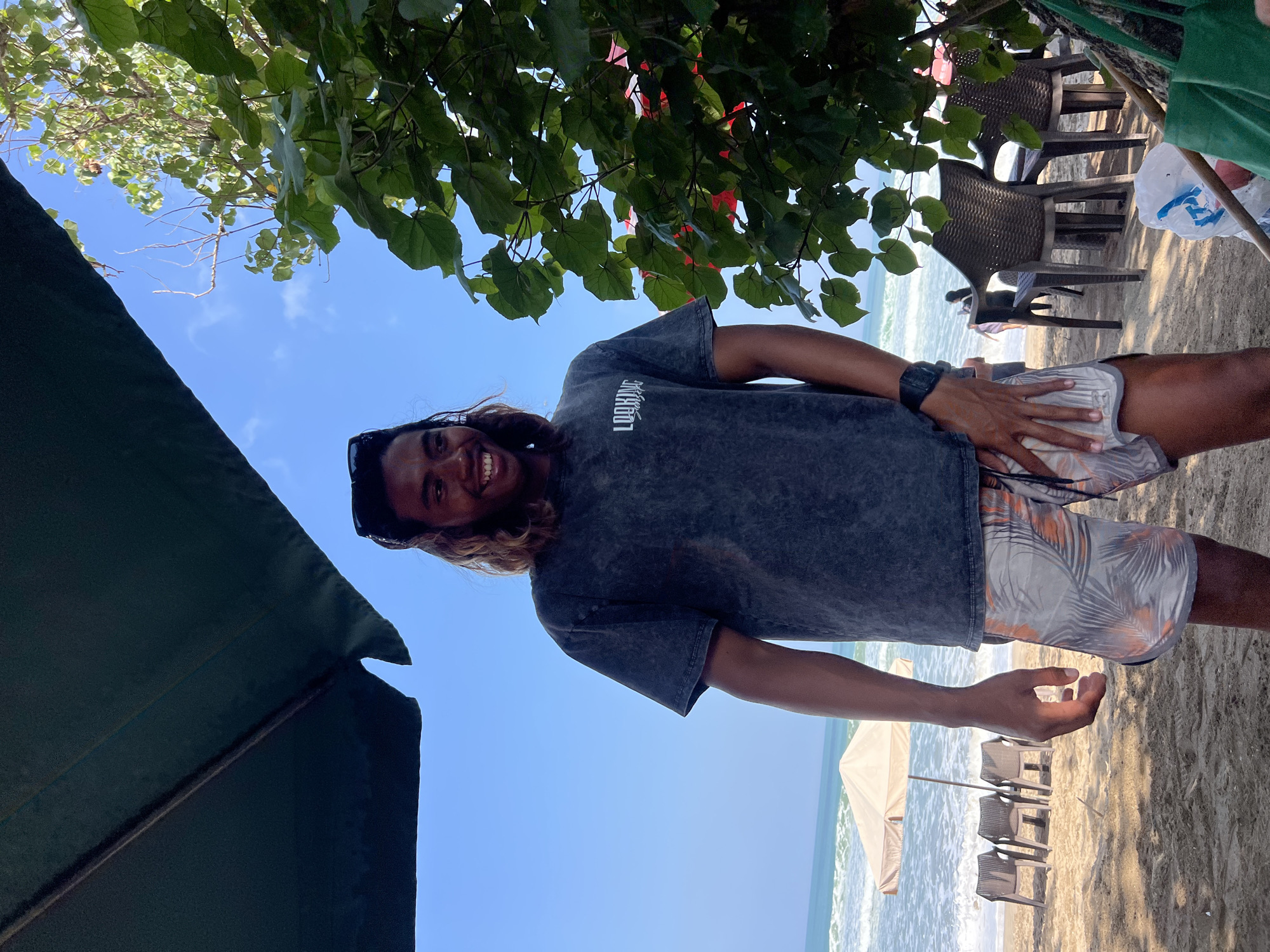 A young Black male with shoulder-length, wavy brown hair and sunglasses perched on his head stands on a sandy beach in Kuta, Indonesia. He is smiling and looking to his right. He wears a dark grey, short-sleeved t-shirt with a white logo on the chest, and patterned grey, orange, and white swim shorts. His left hand rests on his right thigh. A black digital watch is on his left wrist. He is standing under the shade of a large, dark green umbrella, with a lush green tree to his right. In the background, the ocean with moderate waves stretches to the horizon under a clear blue sky with some scattered clouds. Several empty plastic chairs are arranged on the sand, and a light-colored beach umbrella is set up near the water. Other people are visible in the distance, near the water.