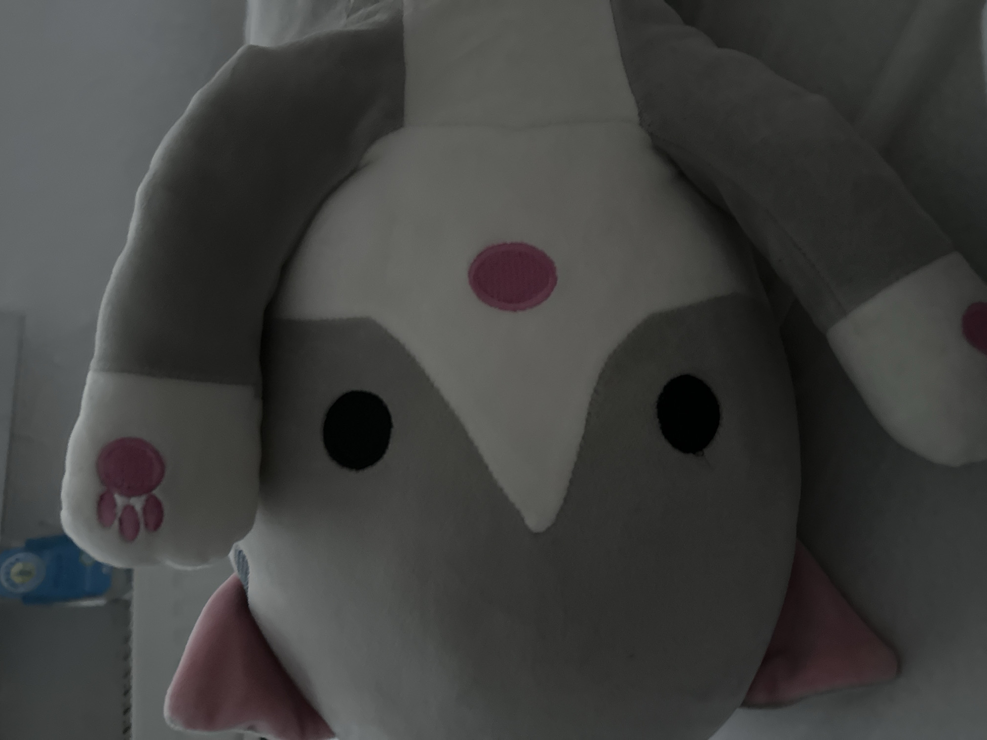 A stuffed cat toy lies on a white surface, possibly a bed. The toy is predominantly gray and white with pink accents on its ears and paw pads. It has black eyes and a pink oval marking on its cheek. The toy appears to be positioned on its side. The background is blurred and shows a blue object, possibly a bottle, on a shelf. There is no indication of the city of Arnstadt, Germany in the image.