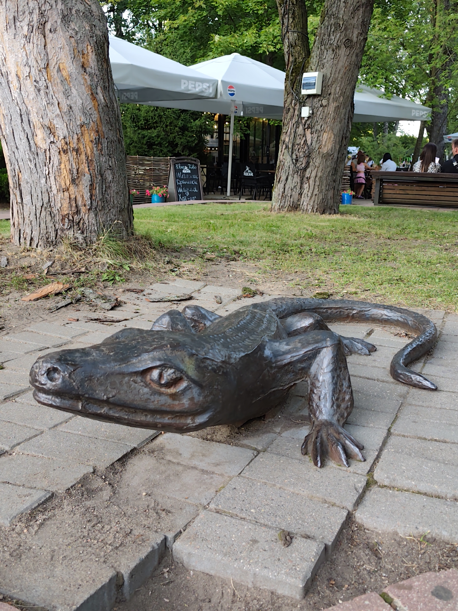 This image depicts a bronze sculpture of a lizard situated on a paved pathway in a park-like setting. The sculpture is the central focus, rendered with intricate detail to mimic scales and limb structure. In the background, there are large trees, a restaurant with outdoor seating under white Pepsi-branded umbrellas, and a grassy area. Several individuals are seated at tables, appearing to be patrons of the restaurant. The ground around the sculpture is a mix of paving stones and soil, with scattered leaves and debris. The overall scene suggests a public outdoor space where art, dining, and nature coexist. Based on visual cues such as the Pepsi branding and the architecture, this location is likely a cafe or restaurant within a park in Minsk, Belarus.