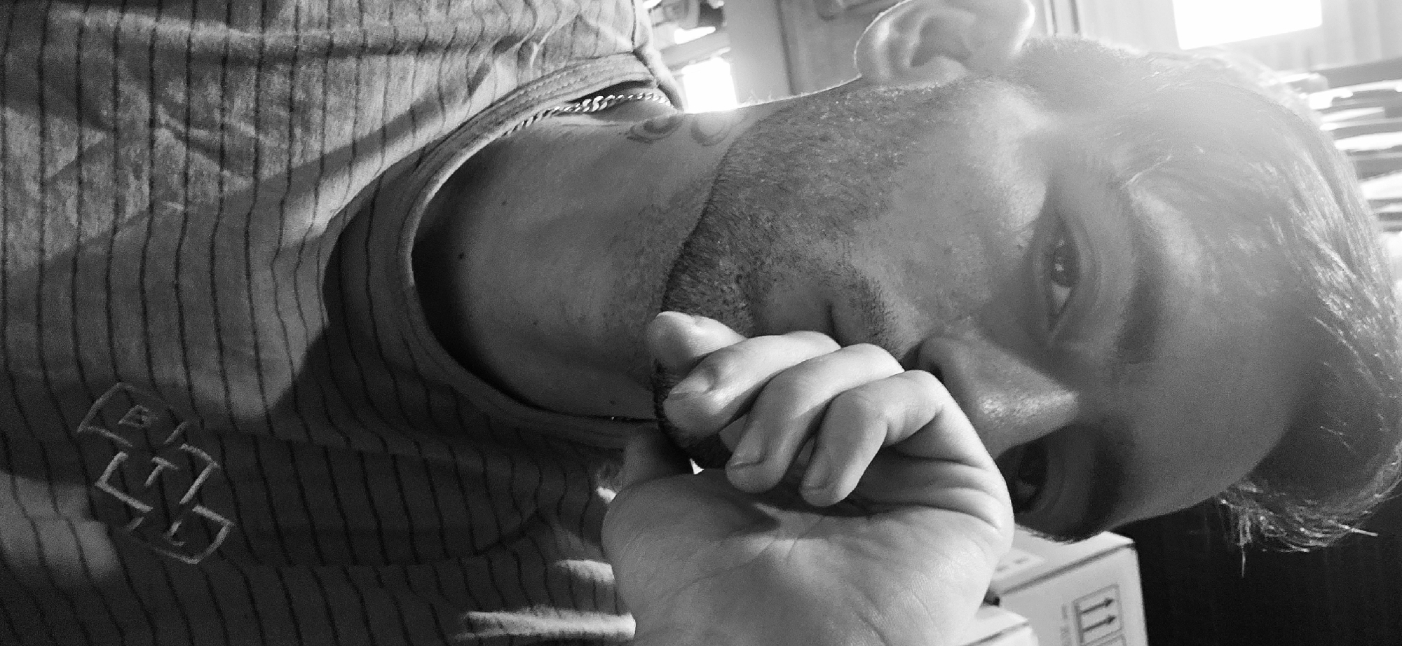 A close-up black and white shot depicts a person, likely male, with a hand near his face, possibly touching his nose or mouth. The person has short hair, a beard, and what appears to be a tattoo on the neck. He's wearing a striped shirt and a chain is visible around his neck. The person's face is partially obscured by his hand. The background is indistinct, but a cardboard box with the number "11" printed on it can be seen, along with papers and shelves in the background. There is no discernible information in this image to specify the location in the city of Plovdiv, Bulgaria. The focus is on the person and the immediate surrounding area.