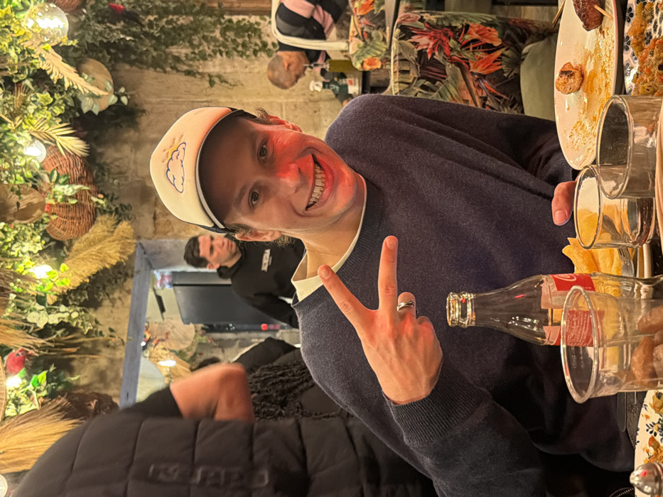 The image depicts the interior of a casual, warm-lit restaurant in Paris, France. A young adult male subject, centered in the foreground, is seated at a table, smiling broadly with teeth showing, and making a peace sign gesture with his left hand. He wears a navy-blue long-sleeved sweater over a lighter-colored collared shirt, a light-colored baseball cap with a blue cloud and yellow sun logo, and a ring on his left ring finger. His face exhibits a reddish hue from ambient lighting.
On the table in front of him are several items: a glass Coca-Cola bottle, multiple clear drinking glasses, a plate containing remnants of food including a partial skewer and a meatball, and another plate holding what appears to be a rice or couscous dish. A small bowl of french fries is also visible.
In the background, other individuals are present. To the left, a person wearing a black puffer vest is partially visible. Directly behind the main subject, another male individual wearing a dark hoodie with a white logo (appearing to be "KUNDRA") is standing. To the far right, an older male subject wearing a pink and white striped shirt is partially visible, seemingly looking down at his table.
The restaurant's decor features rough, light-colored stone or concrete walls. The ceiling is extensively adorned with artificial green foliage, dried grasses, woven baskets, and numerous warm-toned string lights, creating an intimate, naturalistic ambiance. Seating visible in the background includes chairs upholstered with a vibrant tropical floral pattern.
