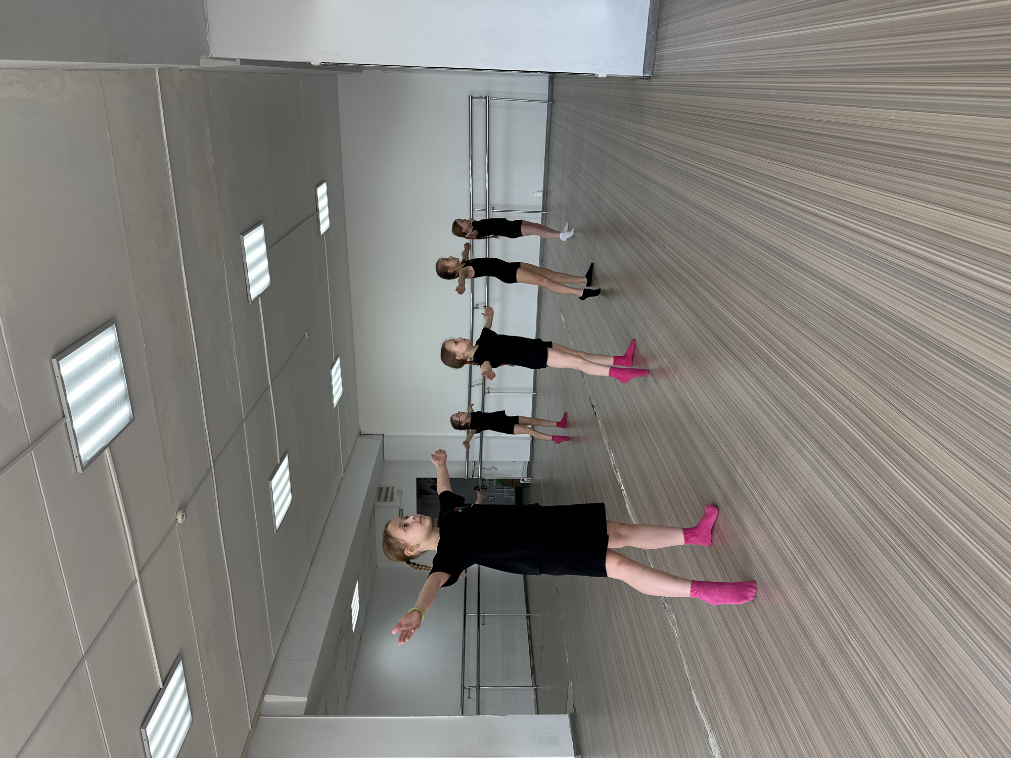 A dance studio in Votkinsk, Russia, has five girls in black dresses and either socks or bare feet standing in a line with their arms out to the sides. The studio has white walls, a grey striped floor, and horizontal metal bars along the wall. The ceiling is white and contains multiple large, rectangular lights.