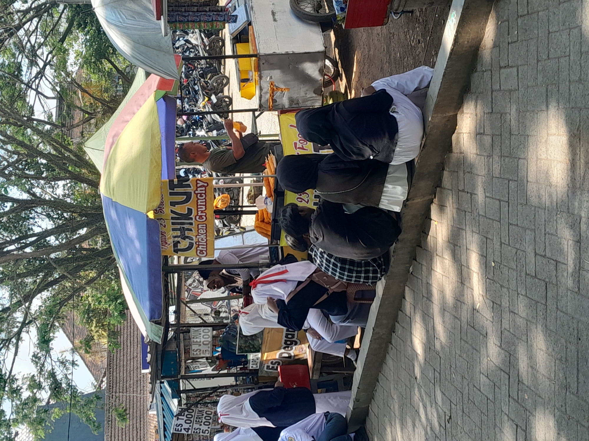 This image depicts a street-side food stall in Genteng Kulon, Indonesia, likely on a sunny day, with dappled sunlight filtering through trees. The stall, adorned with colorful tarpaulins, sells "CHIKUR Chicken Crunchy" and various drinks such as "ES TEH" and "ES JERUK," with prices listed in Indonesian Rupiah (e.g., 4.000, 5.000). A male vendor, wearing a green t-shirt, is actively preparing food, holding a bottle of orange liquid. Several customers, mostly women and young people wearing modest attire including headscarves, are seated on a low concrete barrier in front of the stall, observing the vendor or interacting amongst themselves. Some are facing the stall, while others are looking away. In the background, a line of parked motorcycles suggests a public area or market. The foreground features a paved walkway made of interlocking rectangular stones.