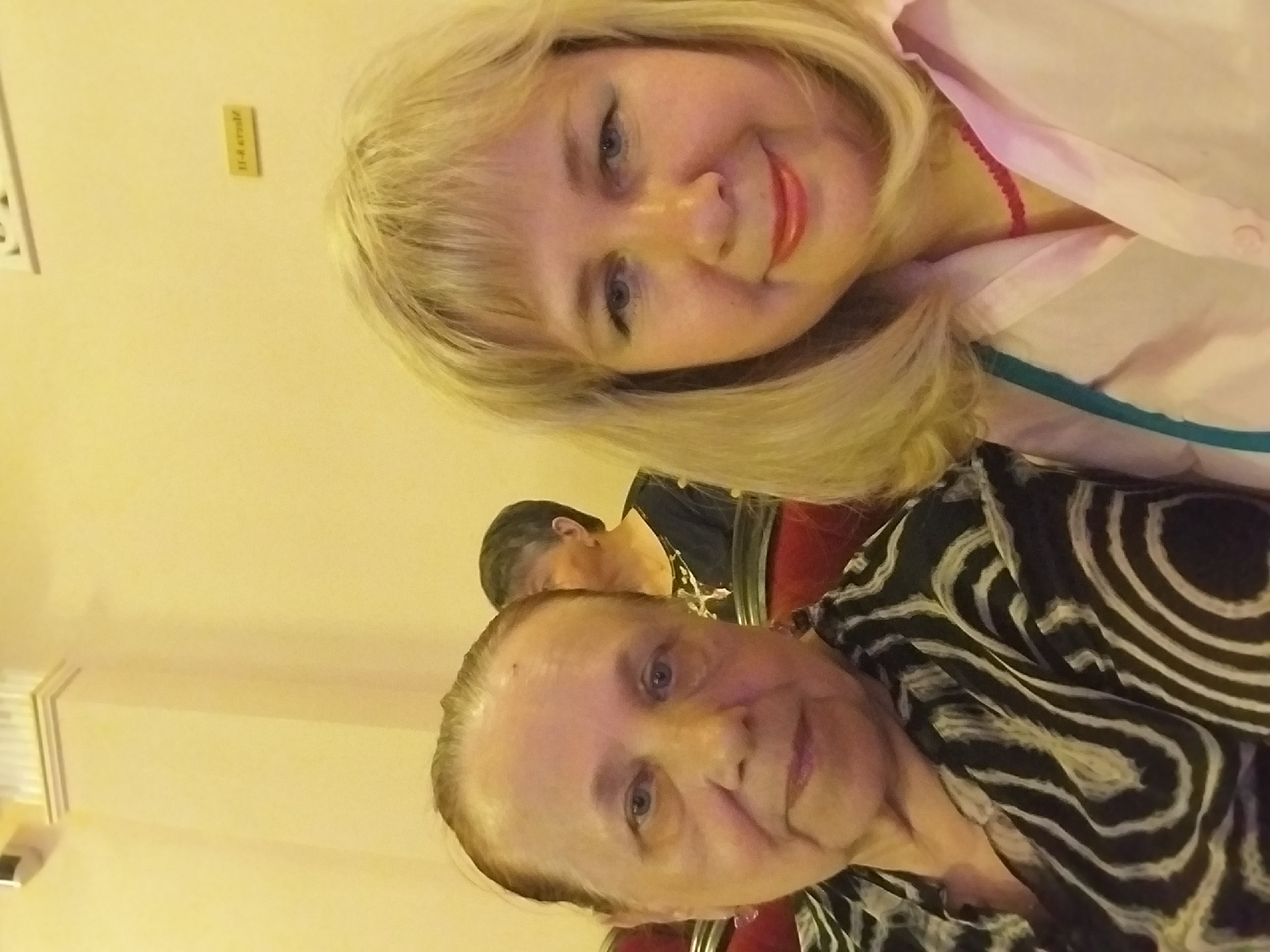 This media file is a selfie taken indoors, likely at an event or gathering, as suggested by the presence of multiple people and seating in the background. The primary focus is on two women. The woman in the foreground, on the right, is older, with fair skin and blue eyes. She is wearing a black and white patterned top. The second woman, positioned behind and to the left of the first, is younger, with blonde hair and blue eyes. She is smiling and wearing a light pink shirt with a red necklace. Another individual is partially visible in the background, with dark hair. The overall lighting is warm, suggesting an indoor setting with artificial lights. A small rectangular sign with text is visible on the wall in the upper left quadrant of the image, but the text is too small to be legible. The image appears to be taken in Orenburg, Russia. The expressions of the subjects suggest a positive and friendly atmosphere.