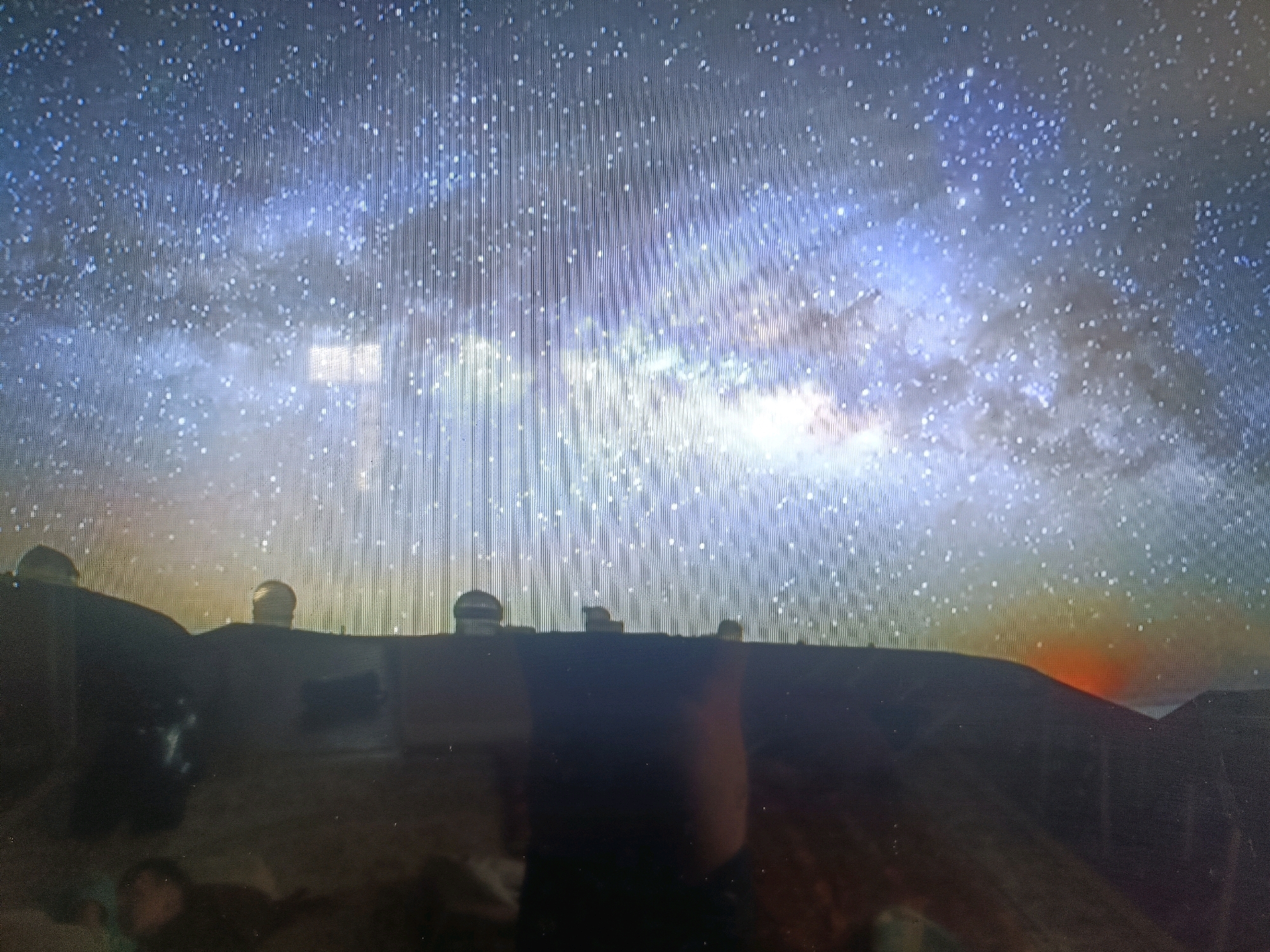 The image depicts a night sky filled with stars, with the Milky Way galaxy prominently visible as a luminous band across the upper portion. In the foreground, silhouetted against the sky, are several domed structures that appear to be observatories or scientific facilities. These structures are situated on a dark, hilly terrain. The lower part of the image is dark and blurry, showing what might be a reflection on a screen or a dimly lit foreground area, with indistinct shapes that could be people. To the right, a faint orange glow on the horizon suggests either a distant light source or the very early stages of sunrise. There is no specific indication or visual element that places this scene within the city of Қасымбек датқа, Kazakhstan.