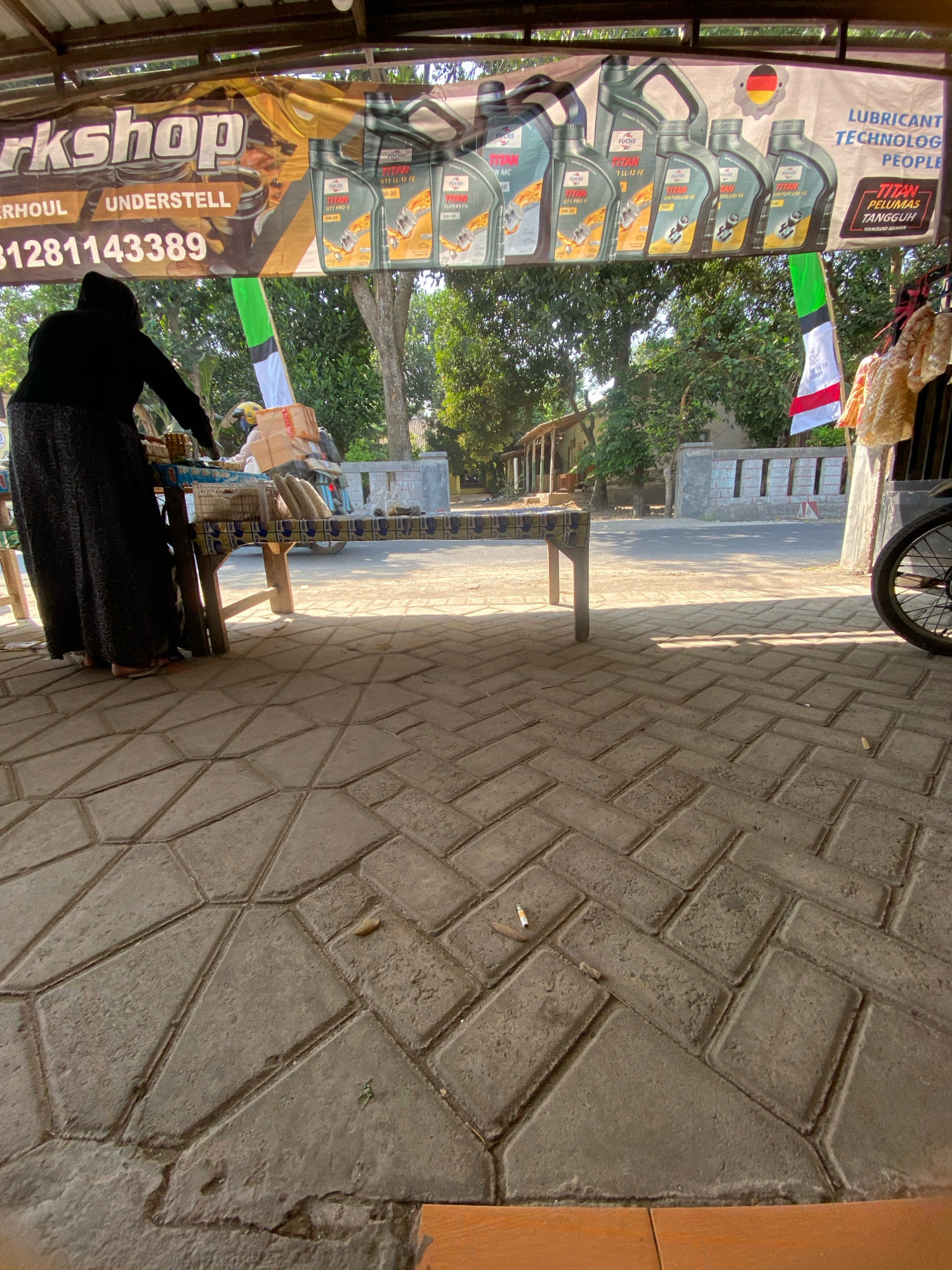 In Gurah, Indonesia, a roadside stall is depicted, possibly a small workshop or convenience store. A woman, partially obscured, stands behind a table displaying various items. In the background, a banner advertises lubricant technology and people. The ground is paved with interlocking concrete blocks, and a cigarette butt rests on the pavement. Trees and buildings line the street, and a motorcycle with boxes stacked on it is visible in the distance. A red, white, and green flag is also present. The scene appears to be outdoors during daylight.
