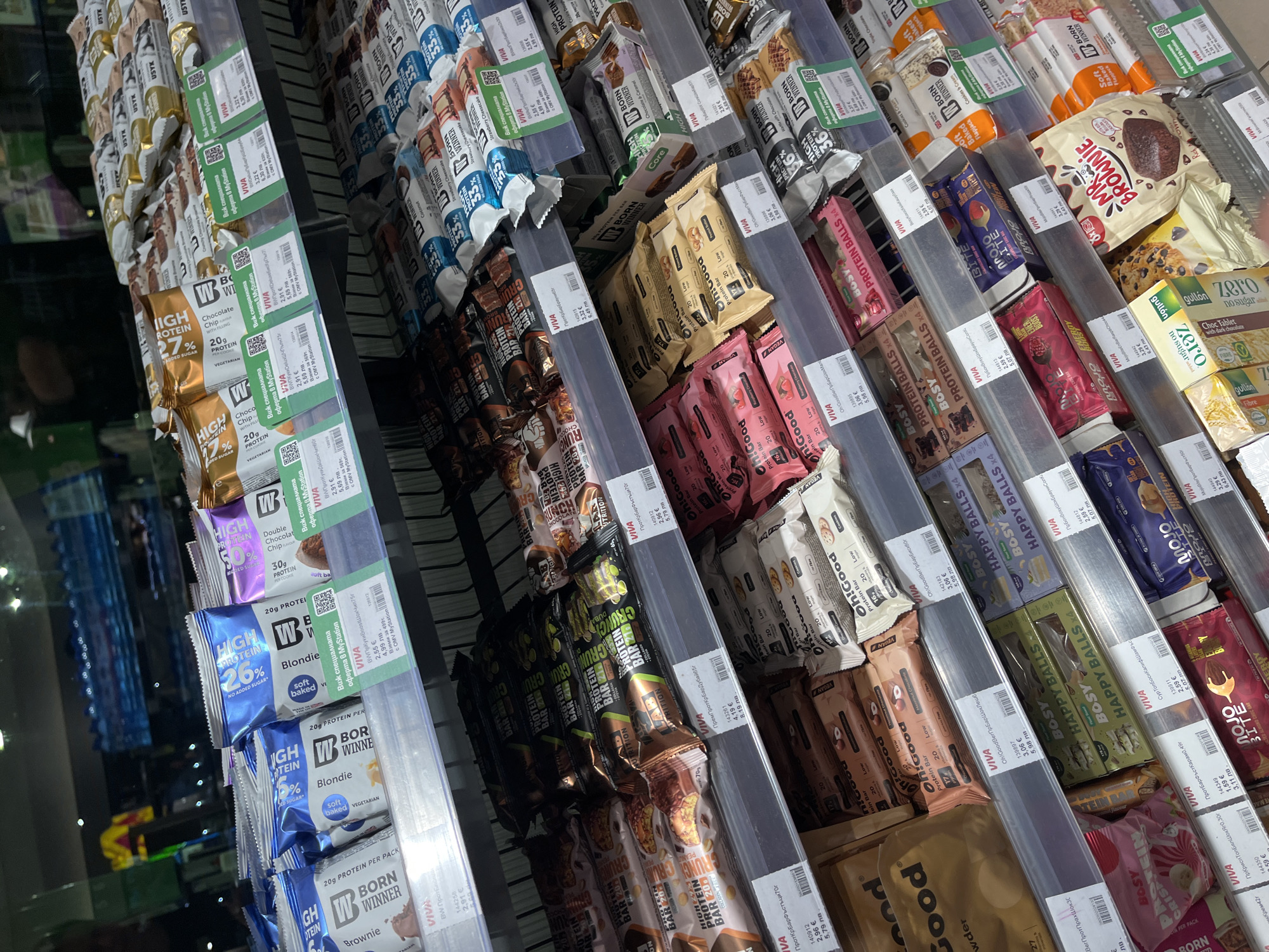An eye-level, slightly angled overhead shot shows shelves of protein bars in a store setting. The protein bars are arranged in clear plastic dividers, and price tags are visible on the front of each divider. The brands of the protein bars include "Born Winner", "W High Protein", "Oh!Good", "Bosy Protein Balls", "Mojo Bite", and "Mr. Brownie". The price tags indicate the prices are in Euros. The location is given on some of the price tags as Popovitsa, Bulgaria, and a store called "Виа".