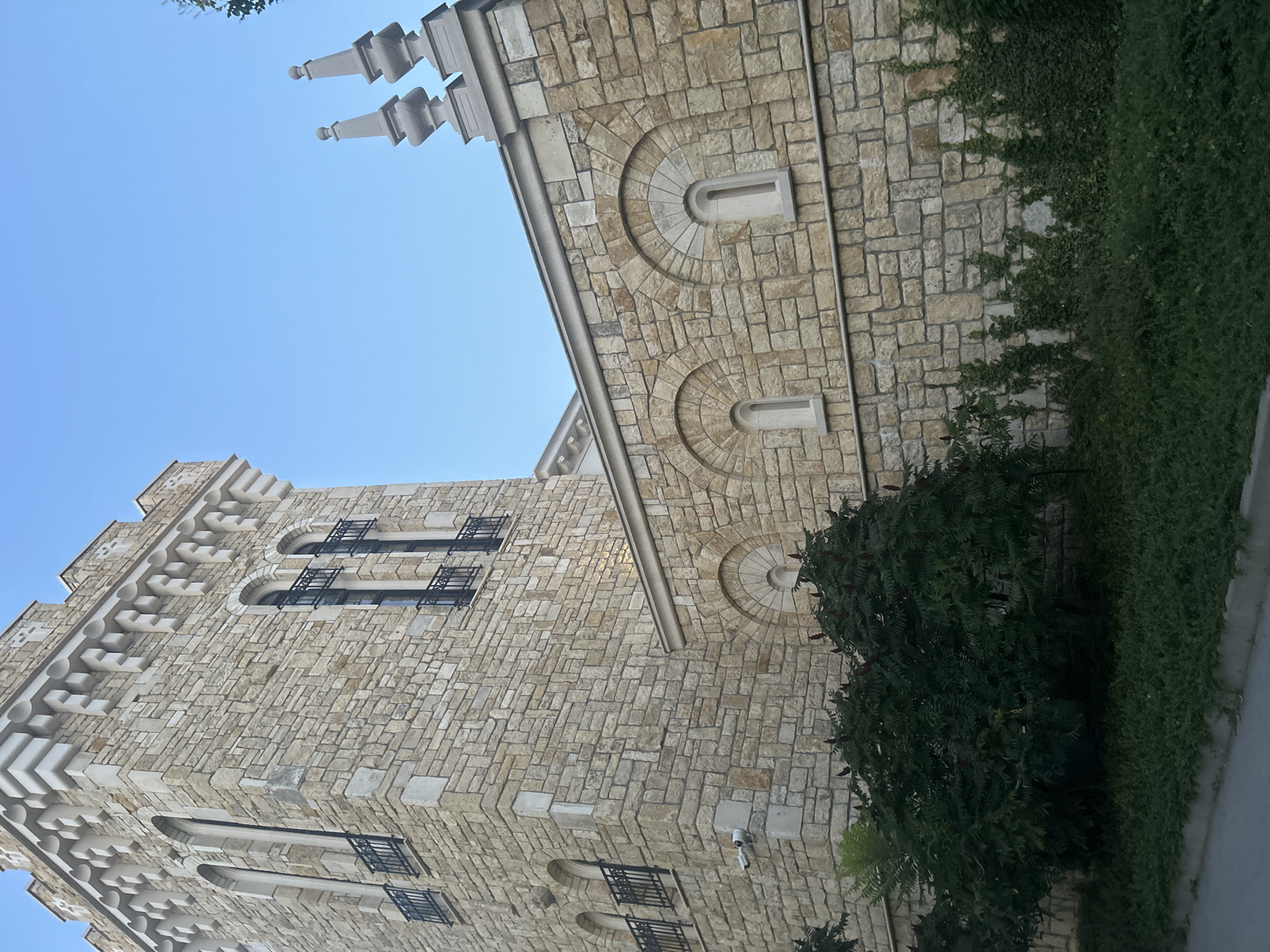 The image shows a building with a stone facade, possibly a church or historical monument, under a clear blue sky. The building features a tower with crenellations and arched windows with black iron railings. Another section of the building has a series of arched window openings. Green foliage and ivy grow around the base of the structure. The architectural style suggests it might be located in the city of Varna, Bulgaria, known for its historical sites. The lighting indicates it is daytime. There are no people or animals visible in the scene.