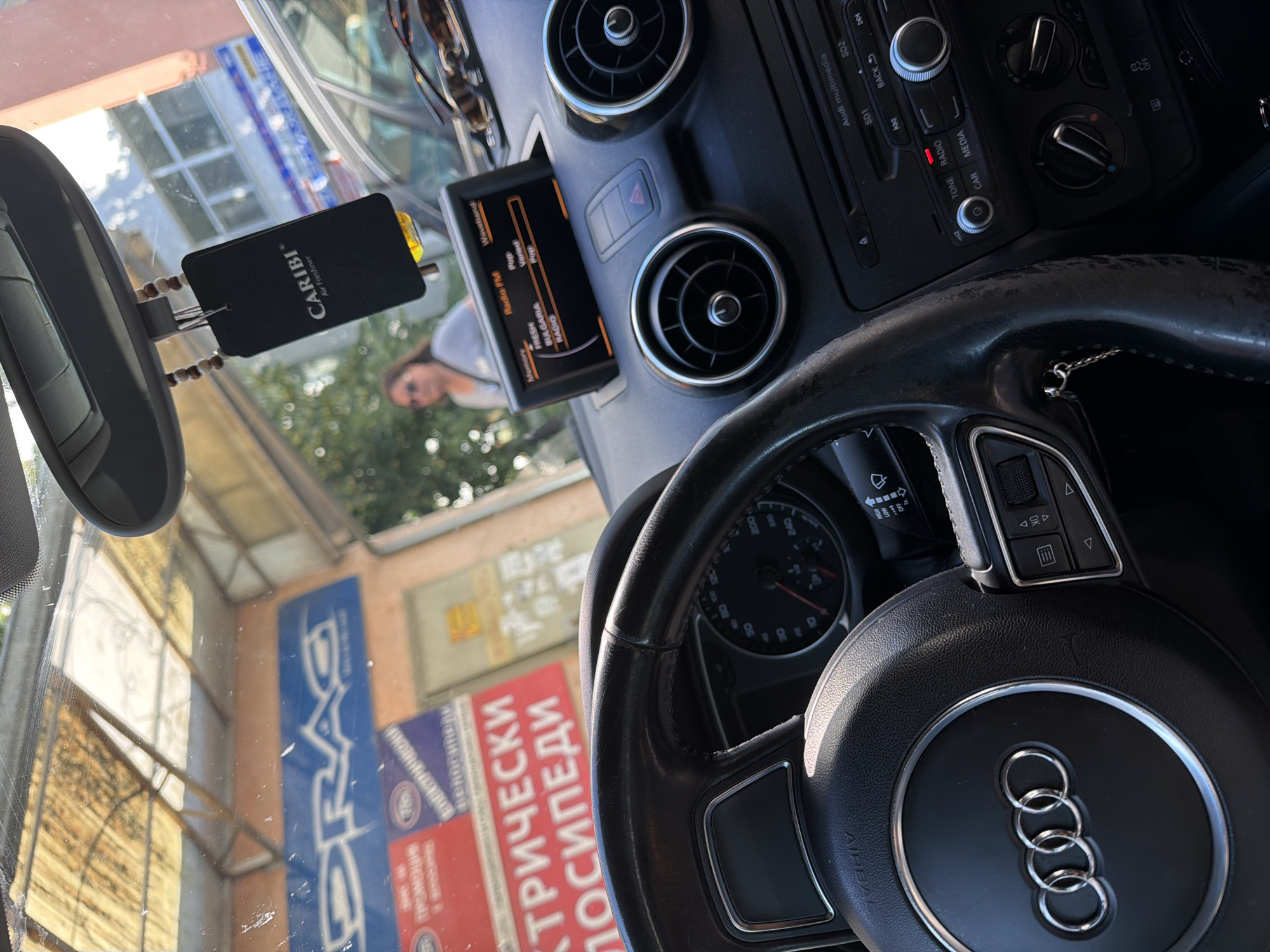 The image shows the interior of an Audi car, looking through the windshield. The steering wheel, dashboard, and infotainment screen are visible. Outside the car, there are signs for "DRAG" and "ELECTRICAL BICYCLES". There is also a person in the background walking. This location is identified as Burgas, Bulgaria, based on the visible text and context.