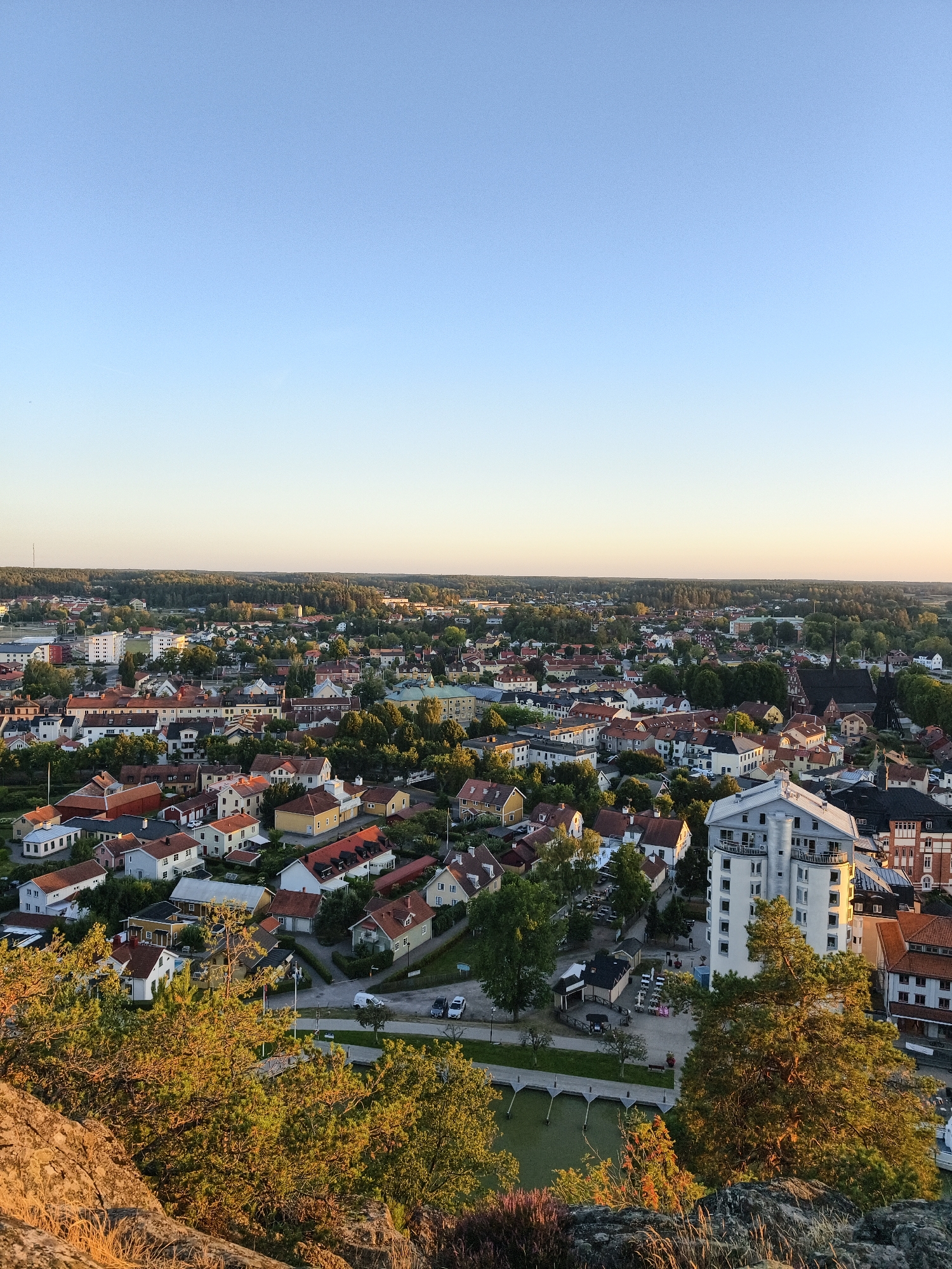 This is an aerial, high-angle, full shot of the city of Söderköping, Sweden, bathed in the warm glow of sunset. The sky above is a clear gradient of light blue to pale yellow, indicating the setting sun. Below, a dense urban landscape unfolds, characterized by a variety of buildings. Red-tiled roofs are prevalent, interspersed with buildings featuring white, yellow, and darker colored facades. Several multi-story apartment buildings and some single-family homes are visible, nestled amongst lush green trees. A prominent tall, white, modern apartment building stands out in the mid-ground. A dark, spire-topped church building is also visible within the city. In the foreground, the rough, rocky texture of a hillside is covered with dry grass and green trees, some of which are illuminated by the low-angled sunlight. A narrow body of water with docked boats and a walkway are present in the lower portion of the frame, adjacent to the residential areas. No people are visible in the image.