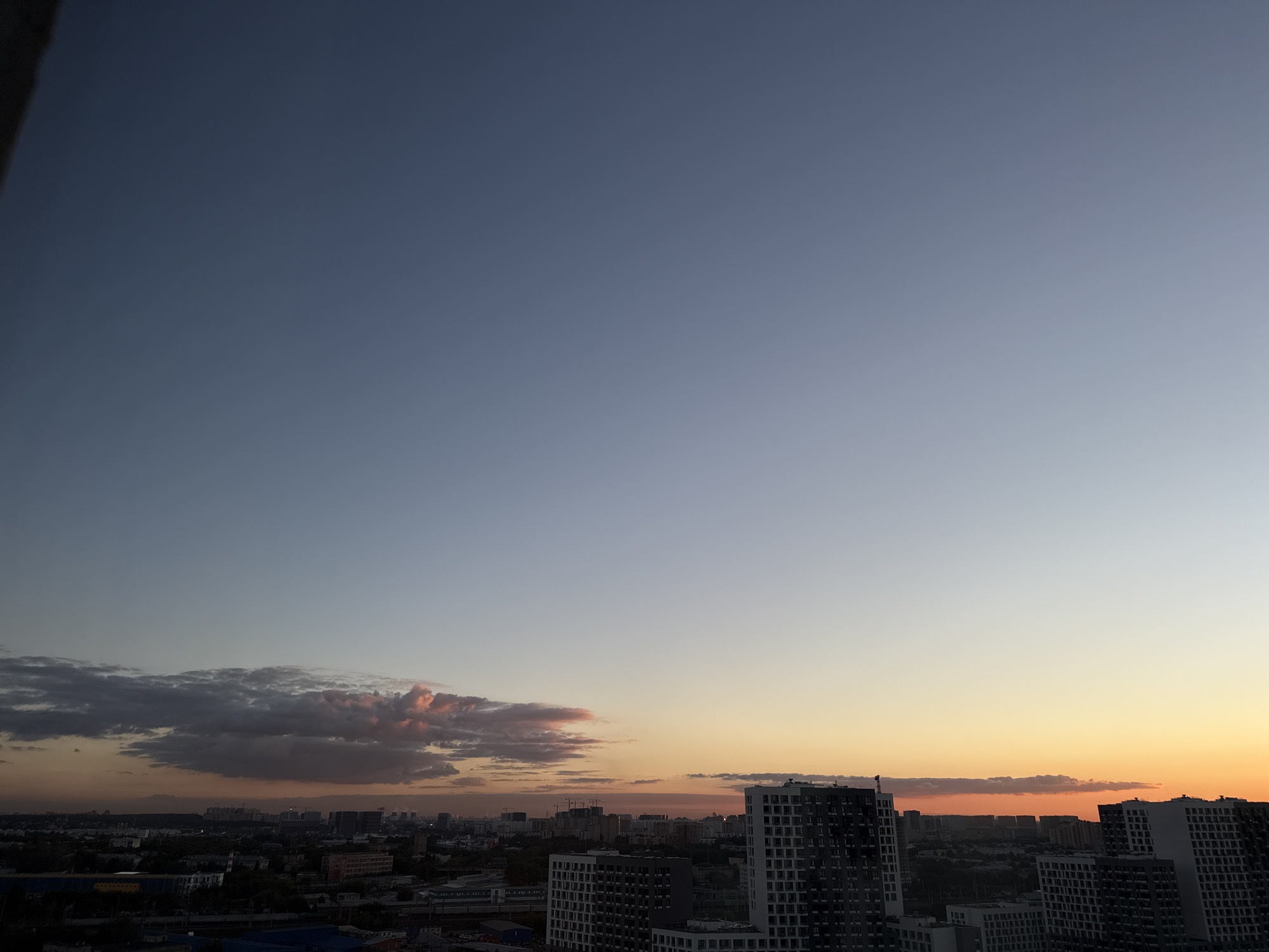 The image captures a panoramic view of Lyubertsy, Russia, during a sunset. The sky transitions from a deep blue at the top to a warm orange and yellow near the horizon, with scattered clouds tinged pink and orange. In the foreground, modern residential buildings with dark and light facades dominate the cityscape. The buildings are tall and appear to be apartments or condominiums, with numerous windows visible. In the background, a lower-lying urban landscape with more varied architecture, including some industrial structures and cranes, stretches towards the horizon. The overall scene depicts a typical evening in a developed urban area, characterized by its high-rise architecture and the atmospheric lighting of sunset. No people are visible in the image.