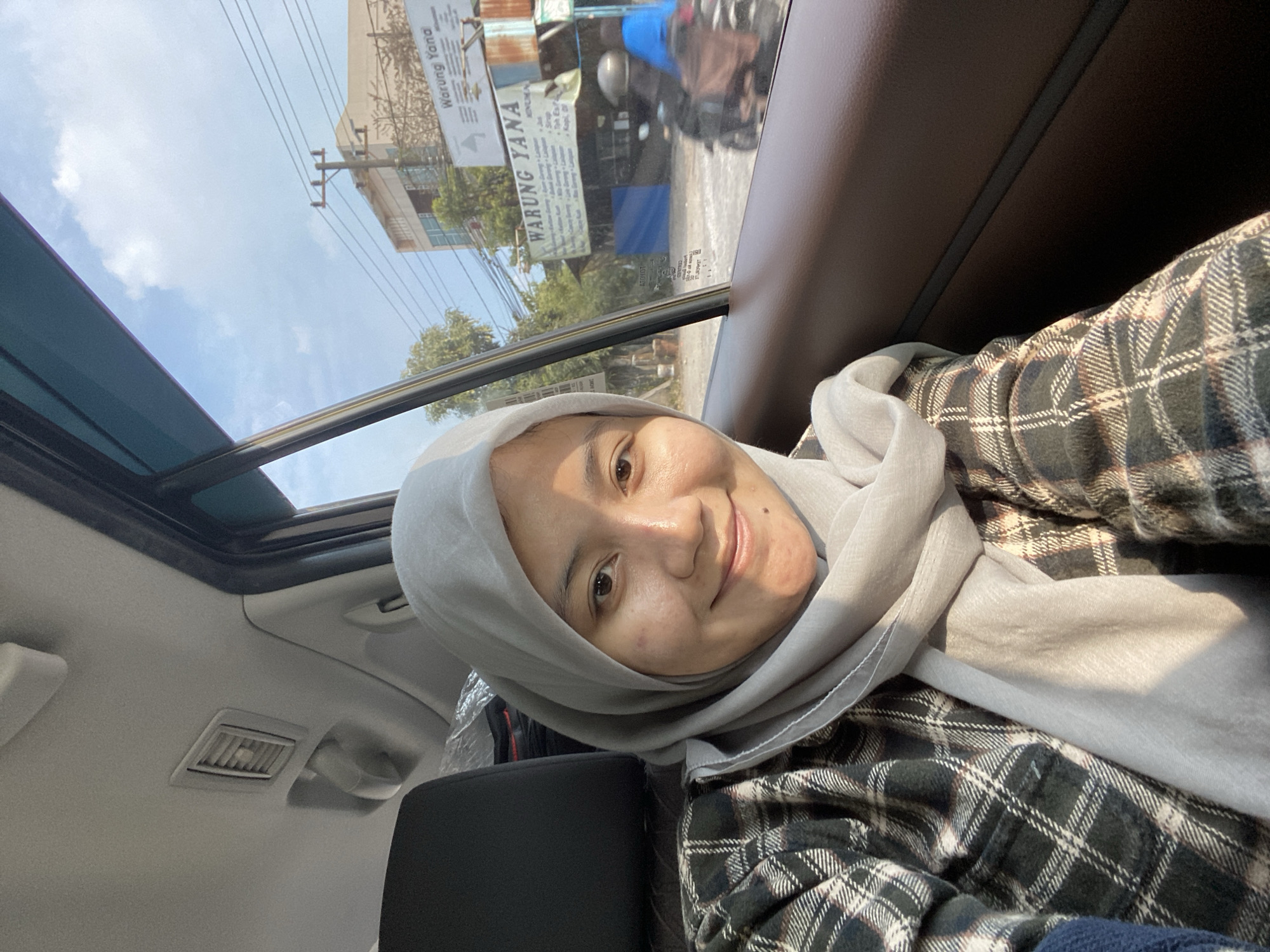 A young Indonesian woman wearing a light grey hijab and a plaid shirt smiles at the camera from inside a car. The car's interior, including a dark brown door panel and a black seat, is visible. Through the car window, a street scene in Gambut Barat, Indonesia, unfolds. A building with signage for "WARUNG YANA" is prominent, alongside power lines and trees. Two people on a motorbike are seen in the background. The sky is blue with scattered clouds, suggesting it is daytime.