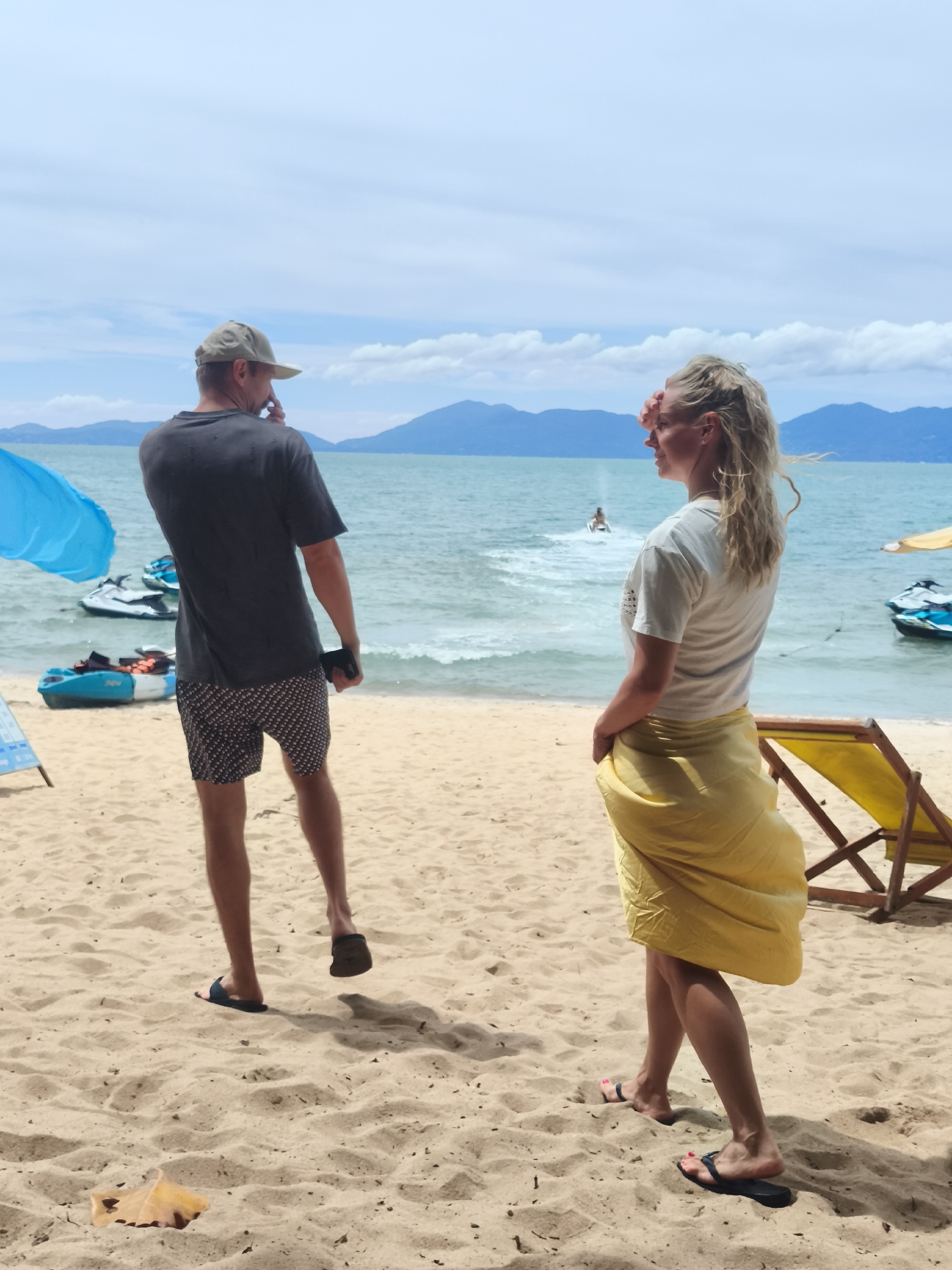 The image depicts a beach scene in Baan Mae Nam, Thailand, under a partly cloudy sky. In the foreground, a man wearing a dark t-shirt, patterned shorts, and a baseball cap stands facing away from the camera, holding his phone to his ear. To his right, a woman with blonde hair, wearing a white t-shirt and a yellow sarong, shields her eyes from the sun as she looks out at the ocean.
The beach is sandy, with a few scattered leaves. In the middle ground, the turquoise ocean stretches towards the horizon, where distant mountains are visible. Several jet skis and a kayak are in the water near the shore. A yellow deck chair is visible on the sand behind the woman. A blue umbrella is partially visible on the left side of the image. The scene appears to capture a moment of leisure and relaxation on a tropical beach.