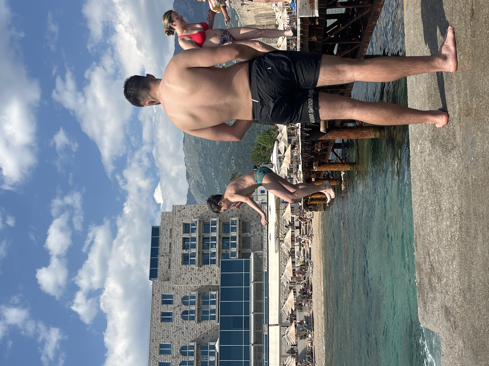 The image shows a group of people at a beach in Budva, Montenegro. The main subjects are a shirtless man standing on a concrete pier, and a boy in green swim trunks mid-jump into the clear blue water. A woman in a red bikini is visible in the background, as are several beach umbrellas and chairs on a sandy beach. The scene is set against a backdrop of mountains and a bright blue sky with fluffy white clouds, suggesting it is a sunny day, likely late morning or early afternoon. The water is calm and transparent, with some ripples visible near the shore. The overall atmosphere appears relaxed and recreational, with people enjoying a summer day at the seaside. The building in the background has a modern architectural style with stone facades and large windows. The text "BOSS BOSS BO" is visible on the man's black swim shorts.