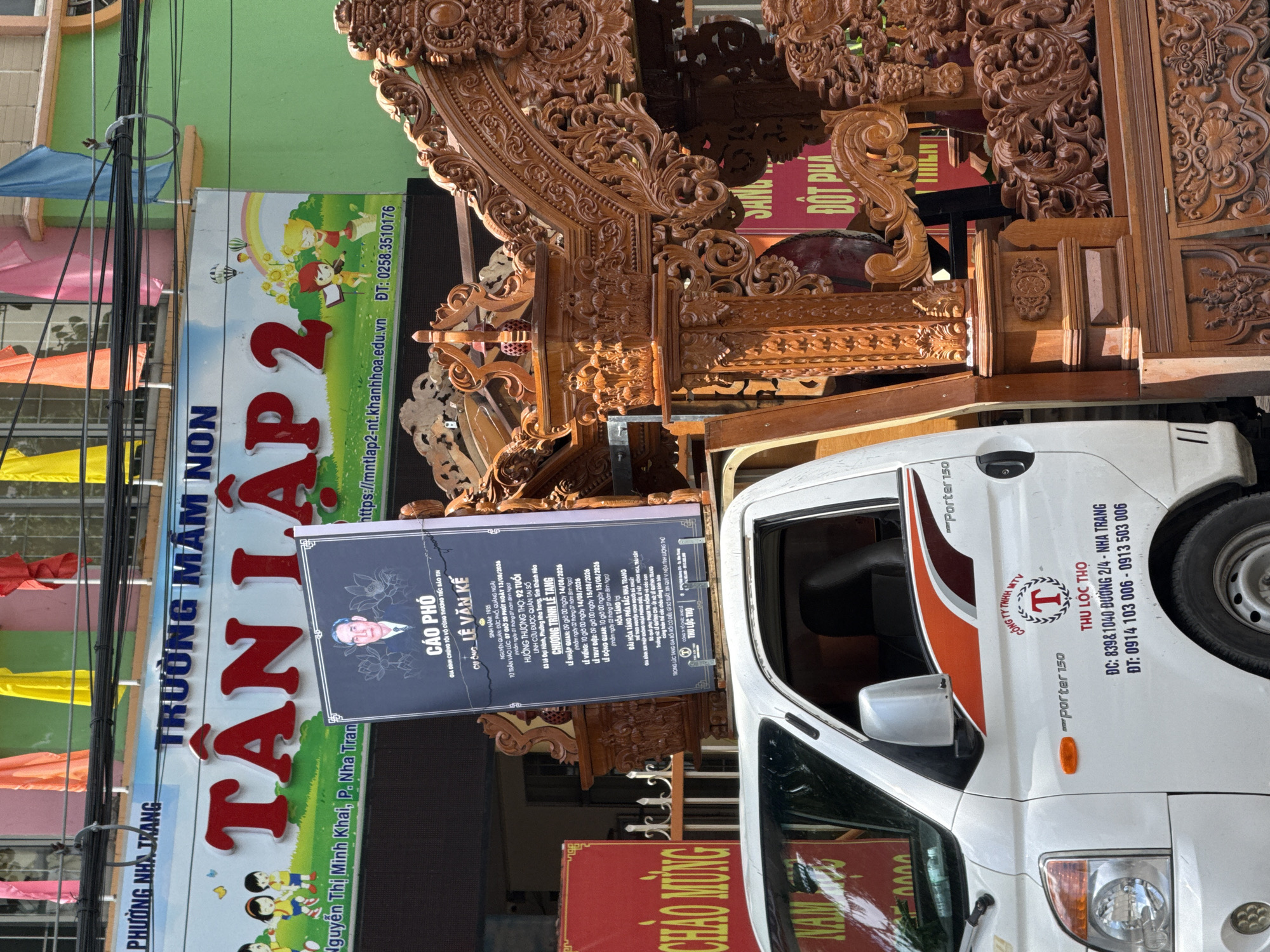 The image shows a funeral procession in Nha Trang, Vietnam. A white truck, labeled "THU LOC THO" and "PORTER 150", is carrying a large, ornate wooden coffin or altar. The truck is parked on a street in front of a building with a sign that reads "TRƯỜNG MẦM NON TÂN LẬP 2" (Tan Lap 2 Kindergarten). A black poster with a portrait of an elderly man is displayed on the truck, bearing funeral announcements. The building's facade is painted green and features colorful flags and banners. In the foreground, a section of an intricately carved wooden structure is visible, likely part of the funeral procession's elaborate decorations. The overall scene suggests a somber event taking place within a residential or educational area of the city.