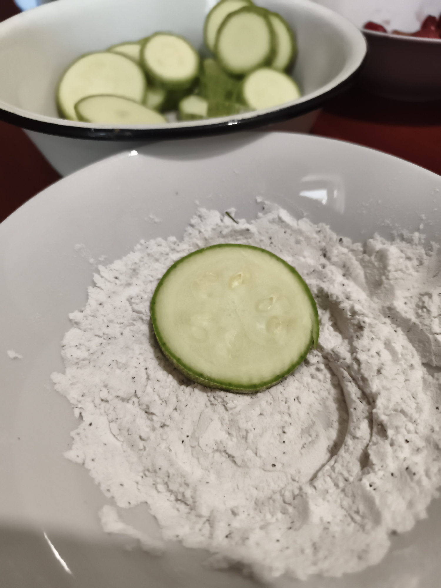 The image shows a close-up view of a white bowl filled with a light-colored powder, possibly flour or a breading mix, with a single slice of zucchini placed on top. The zucchini slice is round with a green rind and a pale green interior, showing the characteristic seeds and texture of the vegetable. In the background, another white bowl is visible, containing more sliced zucchini. The overall scene suggests food preparation, likely for frying or baking. The lighting is natural, indicating daytime, and there are no specific location cues beyond the general context of a kitchen or food preparation area. The mood is neutral, focusing on the ingredients and the early stages of cooking. No text is visible in the image.